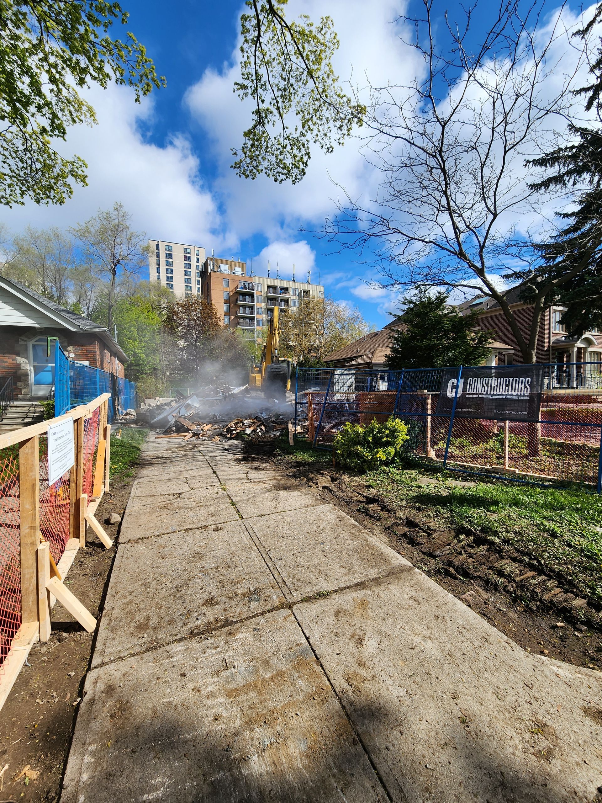 Pathway leading to debris pile with smoke, construction in background; trees and building under a cloudy sky.