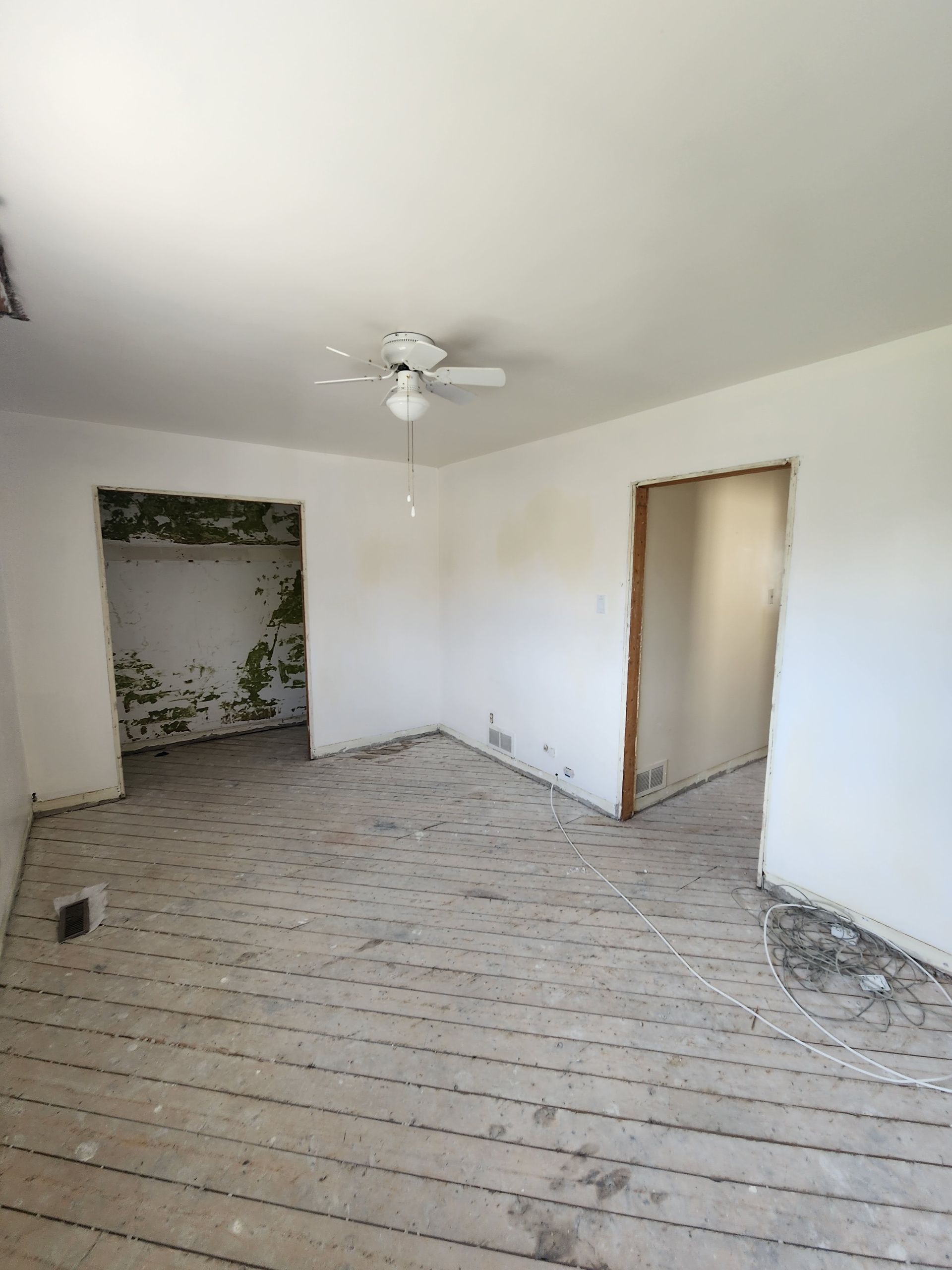 Empty room with exposed subfloor, two doorways, and a ceiling fan. Walls are painted white.