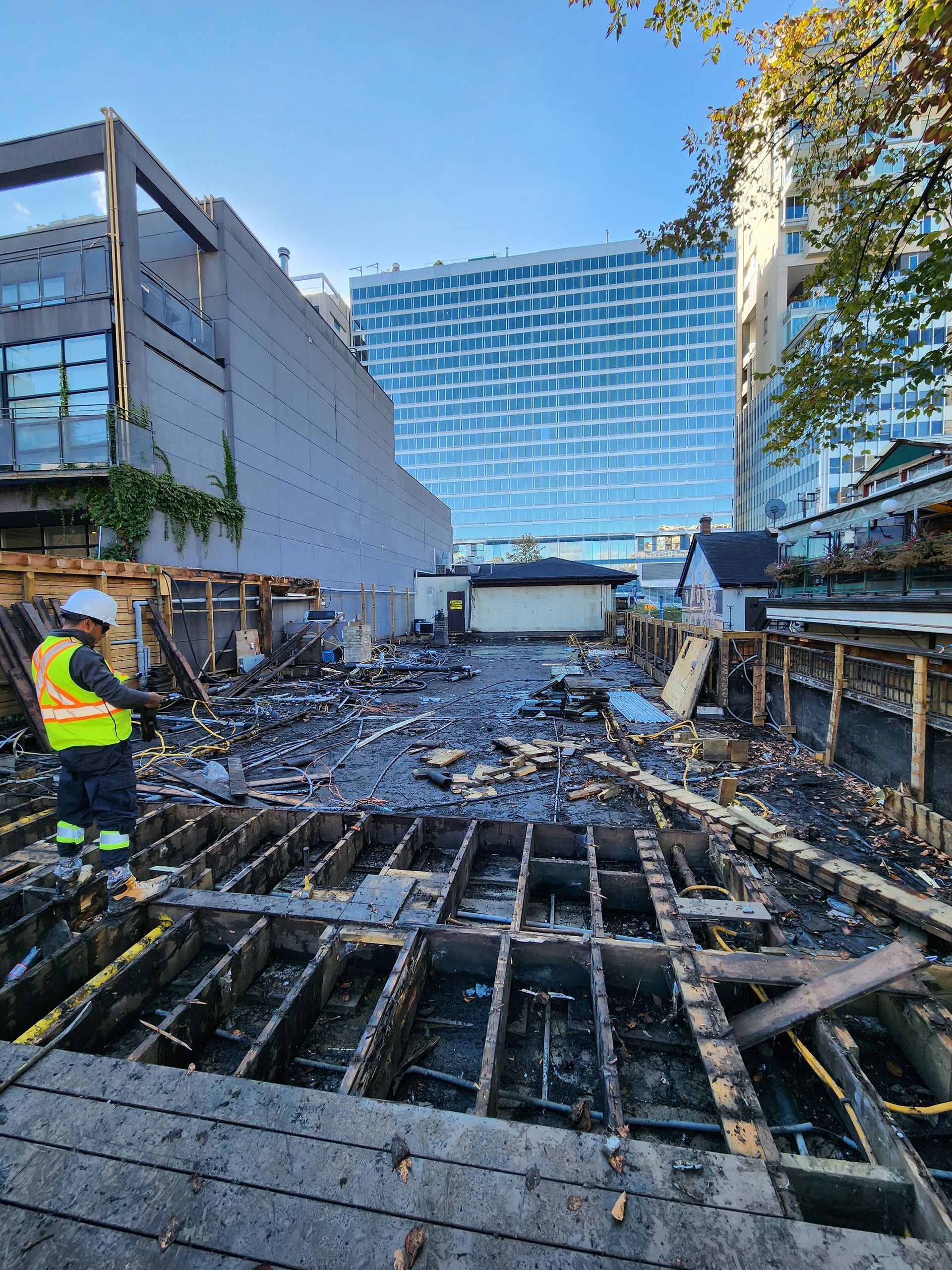 Construction site with worker in vest, debris, and buildings in background.