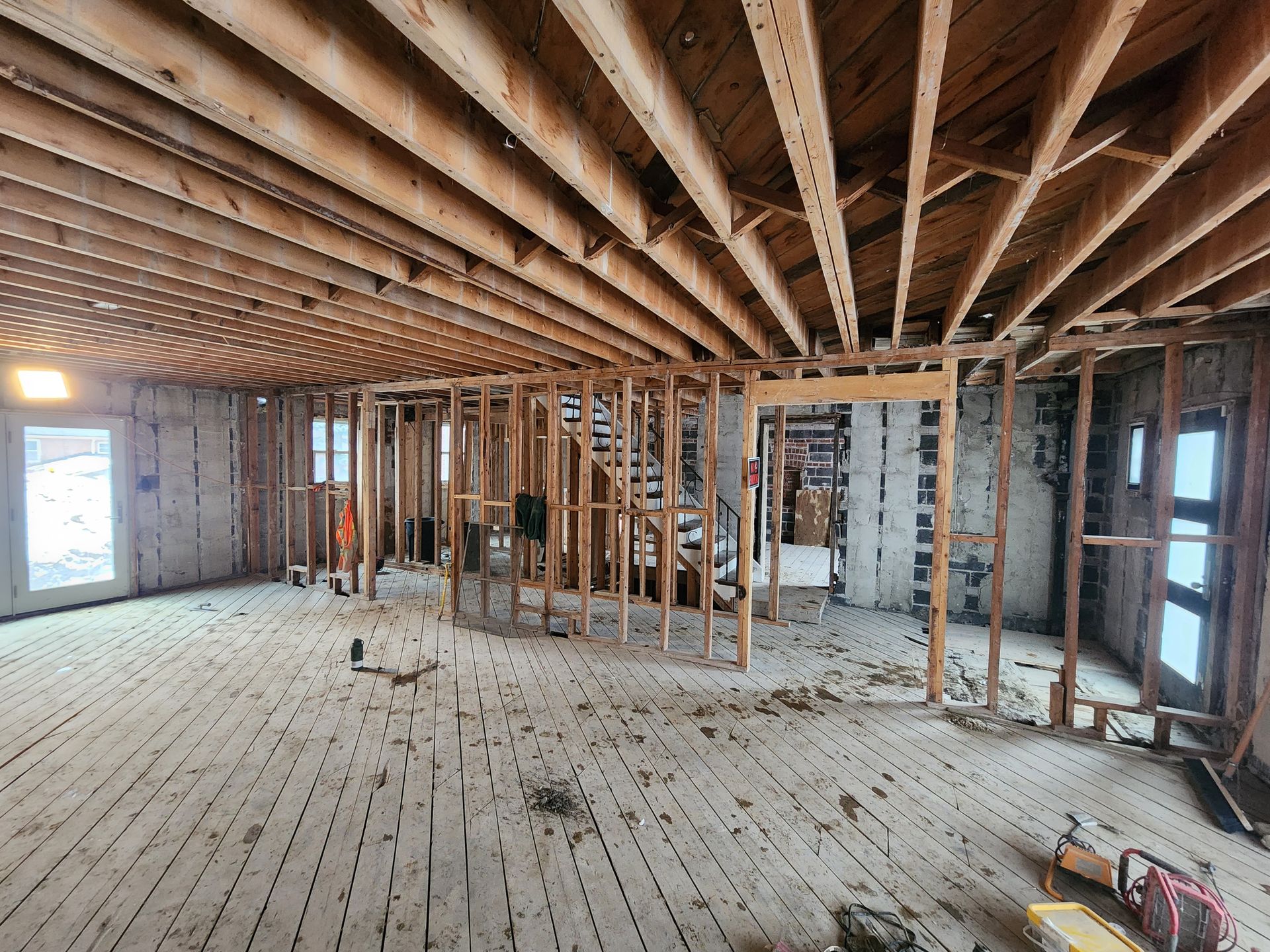 Interior undergoing renovation, exposed wood framing, ceiling joists, and flooring. Natural light streams in.