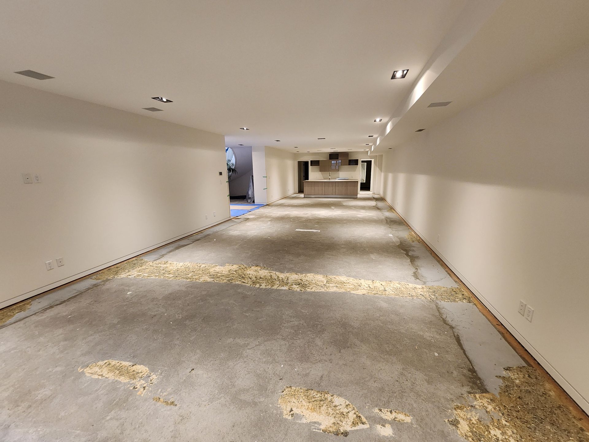 Empty, unfinished basement with debris on the concrete floor. White walls, recessed lighting, and kitchen visible in the distance.