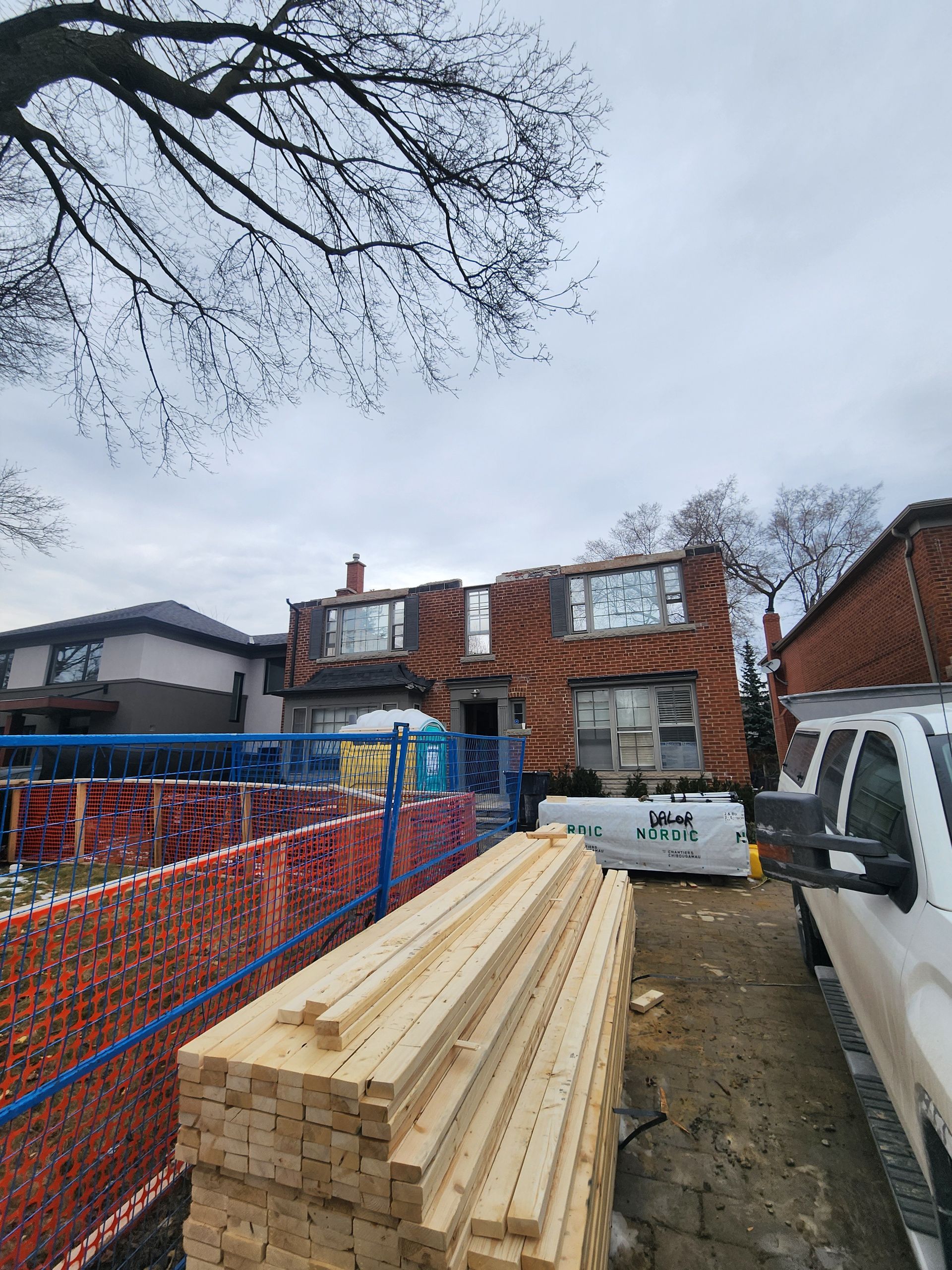House under construction with stacks of lumber in the foreground; blue construction fence, cloudy sky.