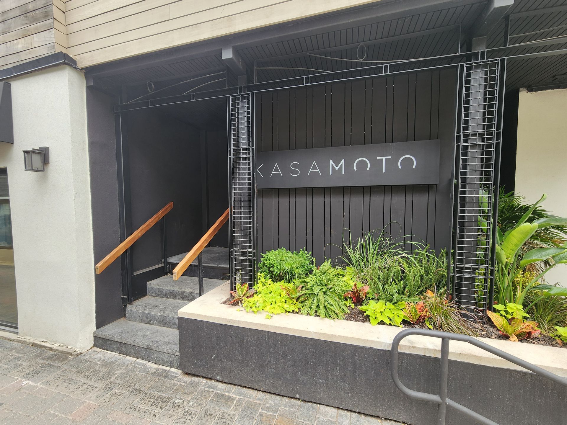Entrance to Kasamoto restaurant with black facade, sign, steps, and greenery.