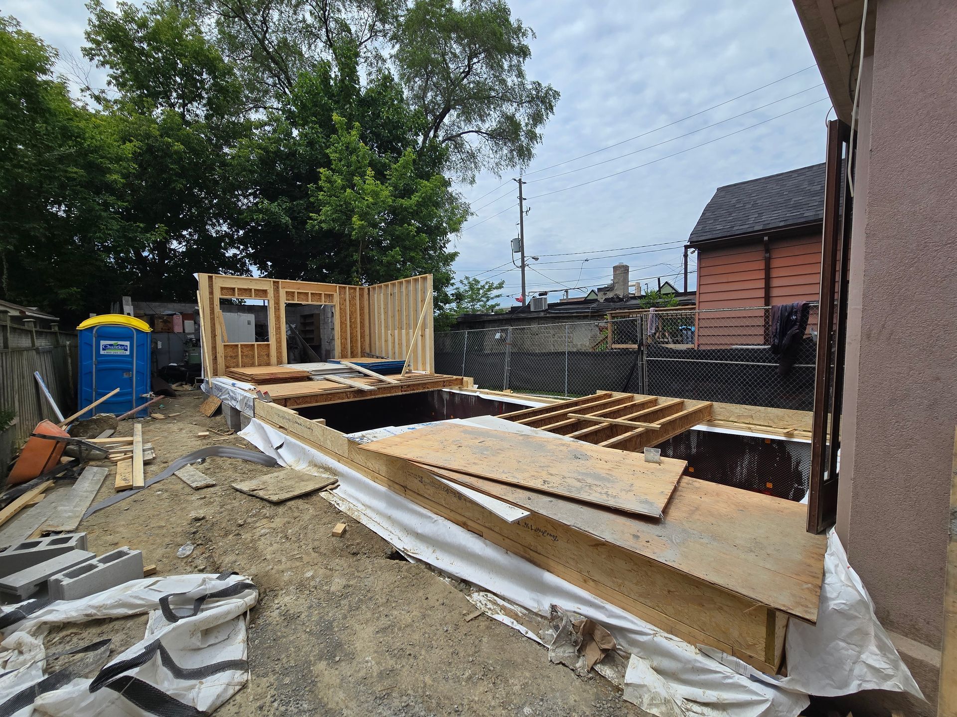 Construction site: wooden structure framing, decking, materials. Blue portable toilet, brick building in background.