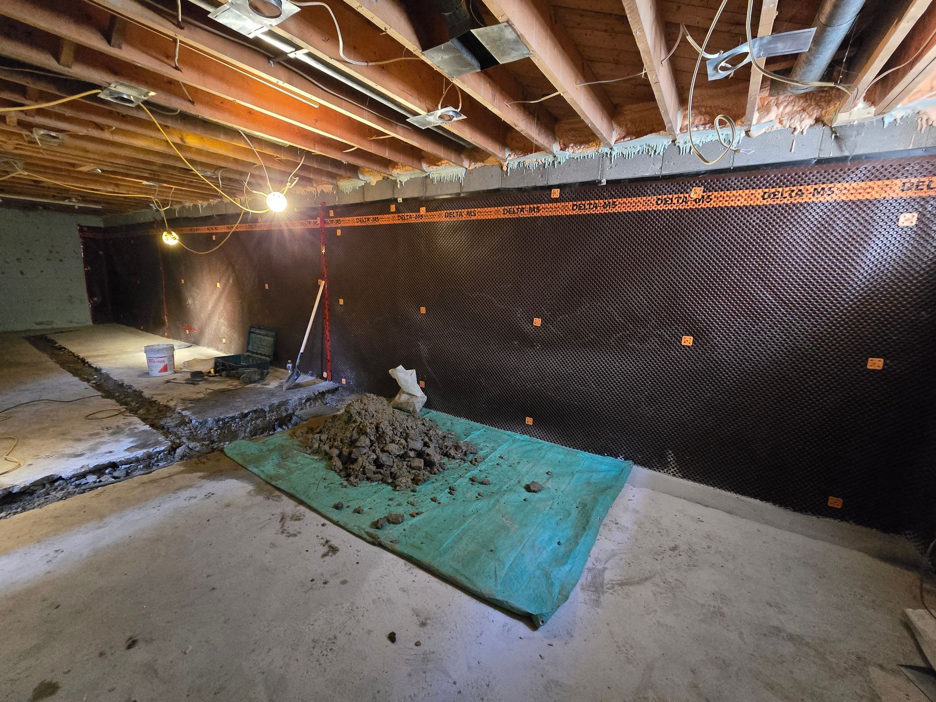 Basement renovation in progress. Black dimpled drainage membrane covers a wall. Ground has concrete and a green tarp with dirt.
