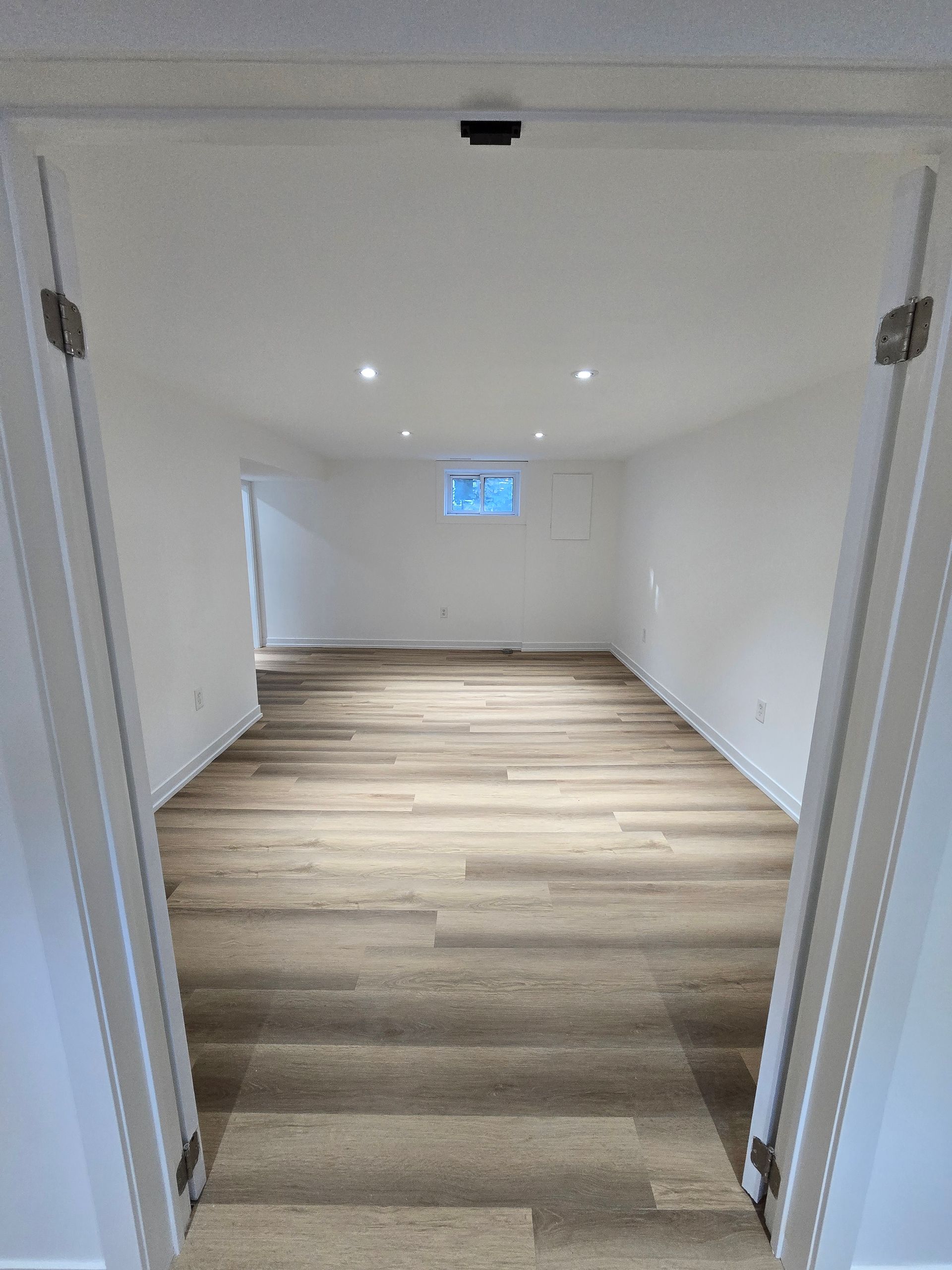 Empty, rectangular room with light-colored flooring, white walls, recessed lighting, and a small window.