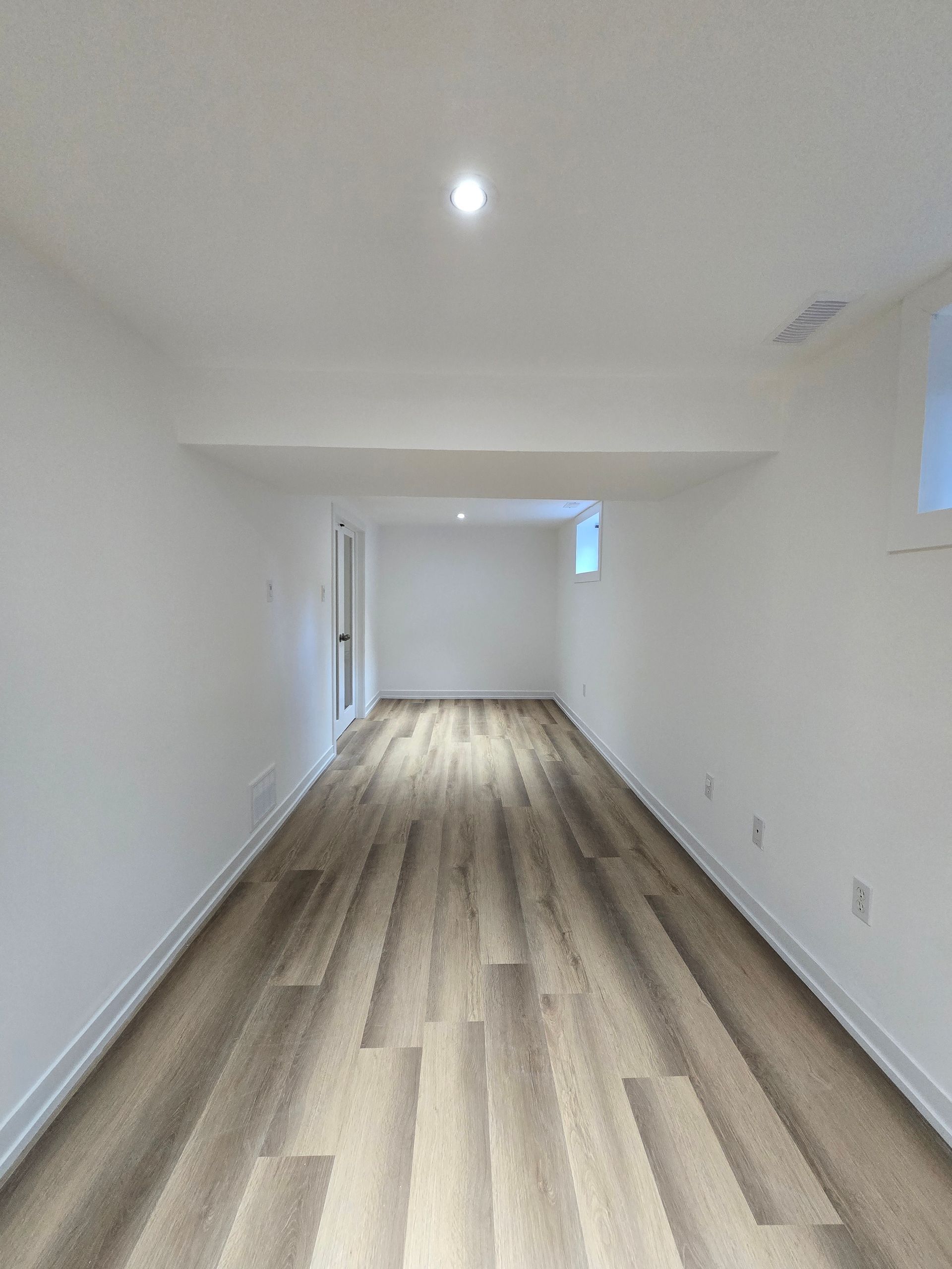 Empty, long, rectangular room. White walls, wood-look flooring. Recessed ceiling lights.
