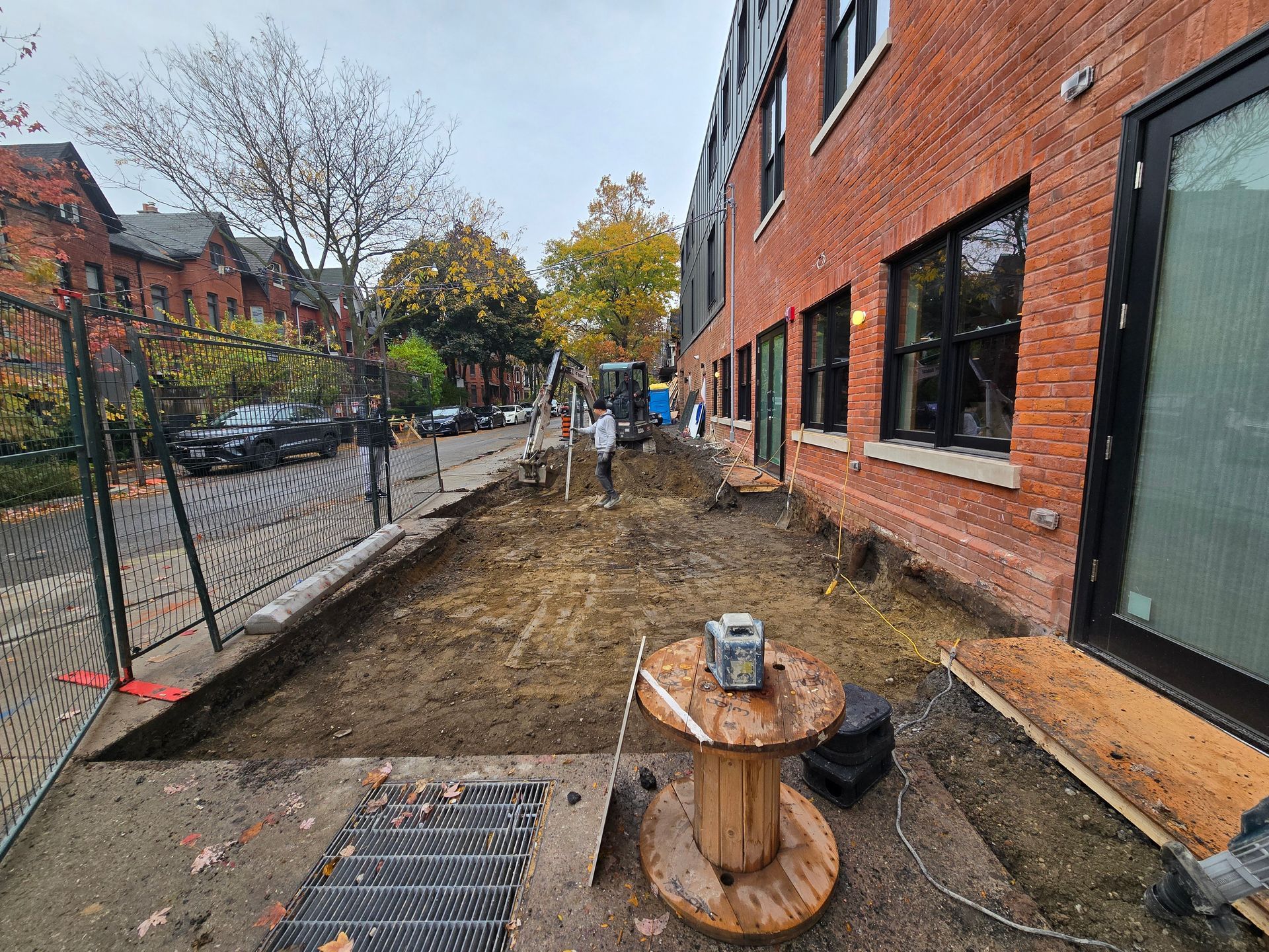 Construction site with exposed earth next to a brick building. Trees and parked cars are visible on the street.