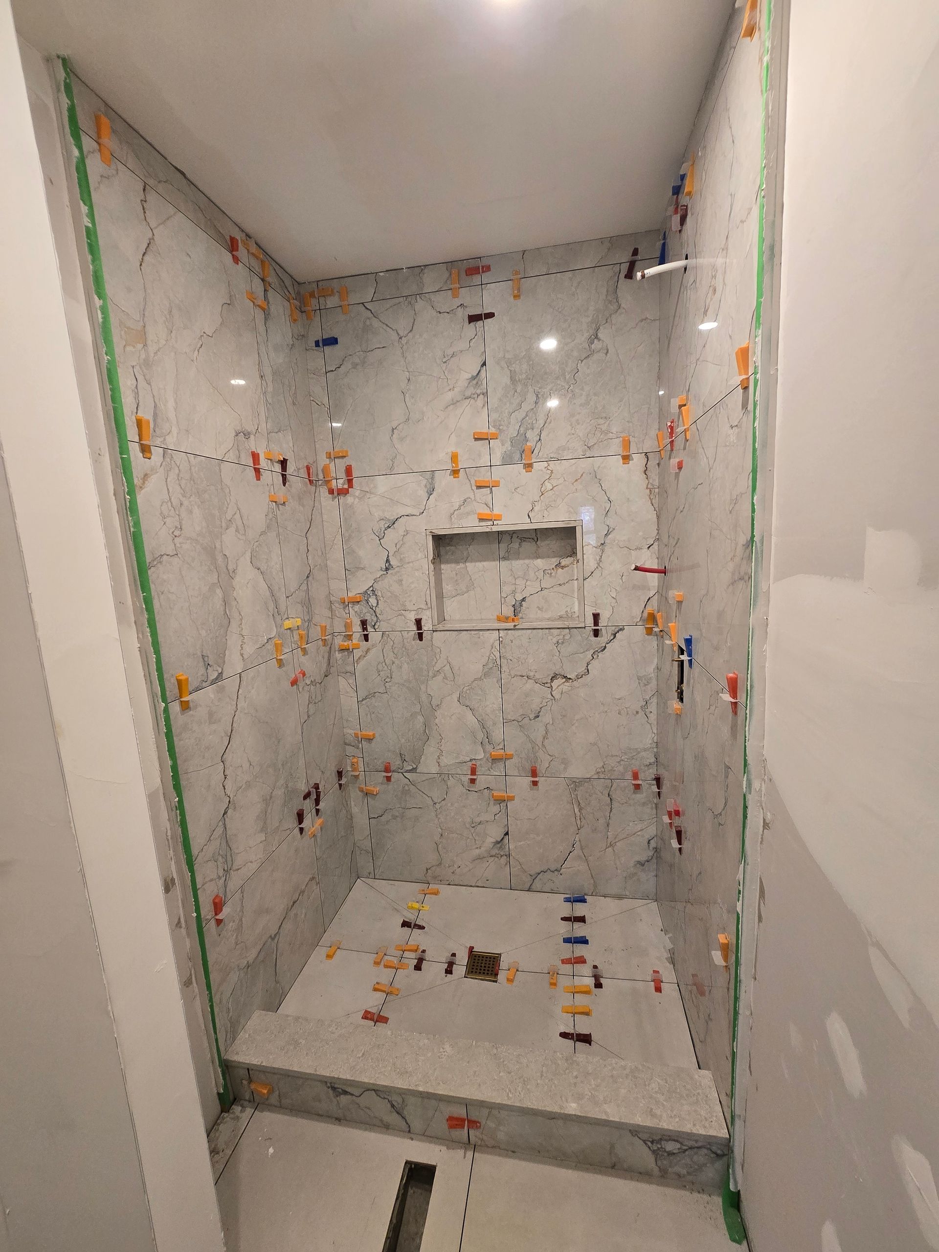 Tiled shower with gray marble-look tiles, a built-in niche, and orange leveling clips in a white room.
