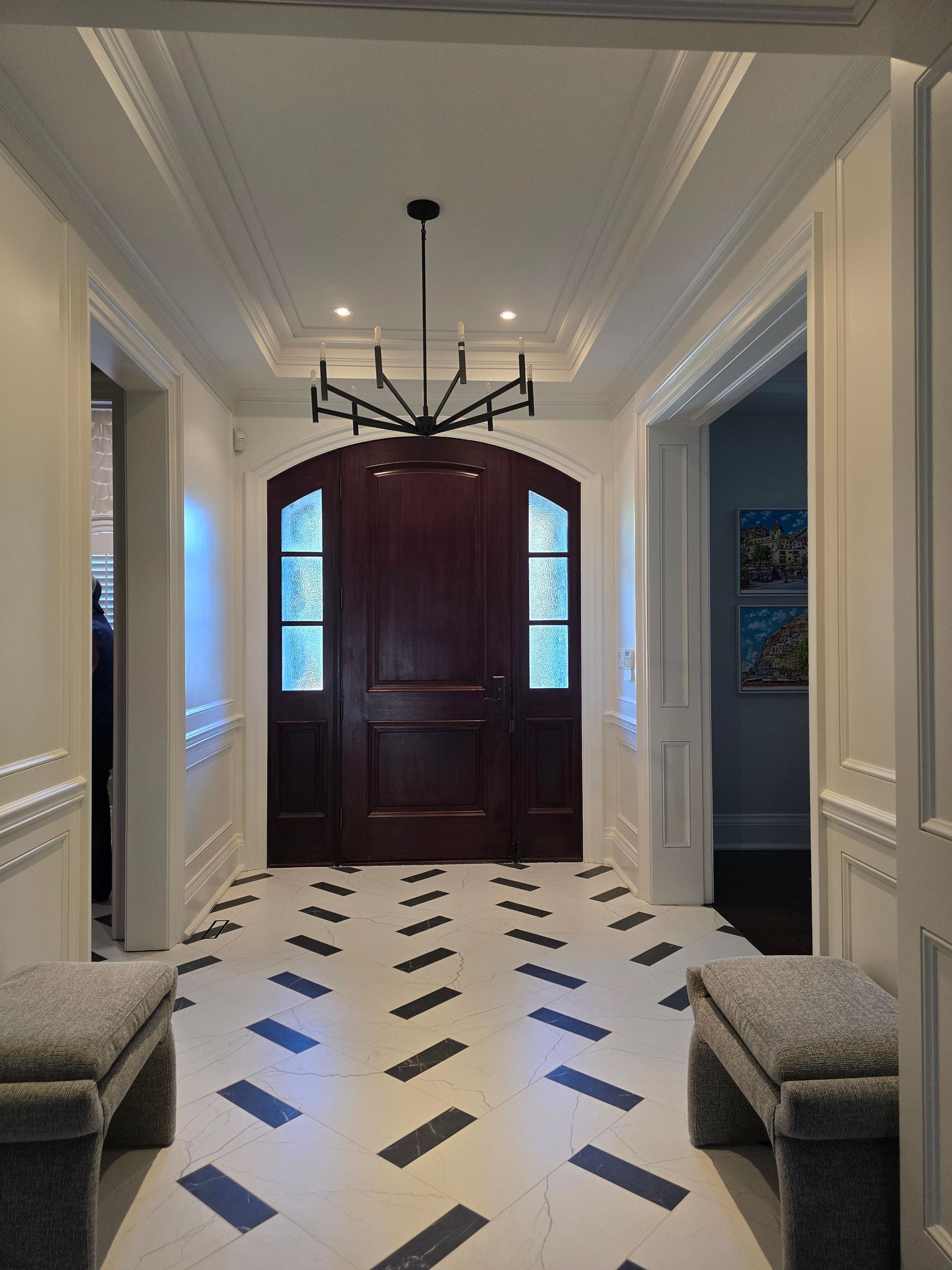 Elegant hallway with dark wood door, patterned floor, two benches, and chandelier.