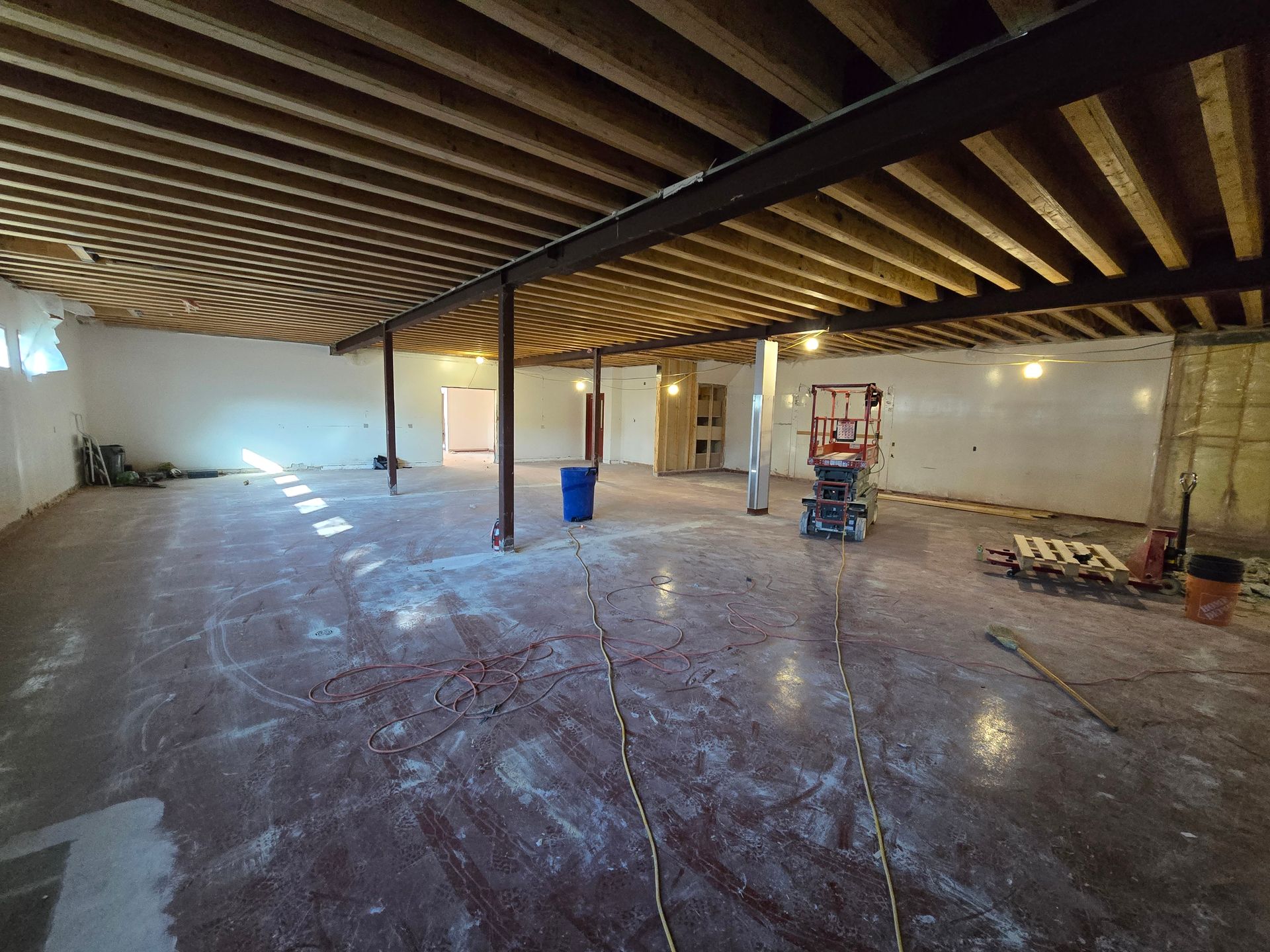 Renovated basement interior. Concrete floor with exposed joists, steel beams, support columns, and construction equipment.