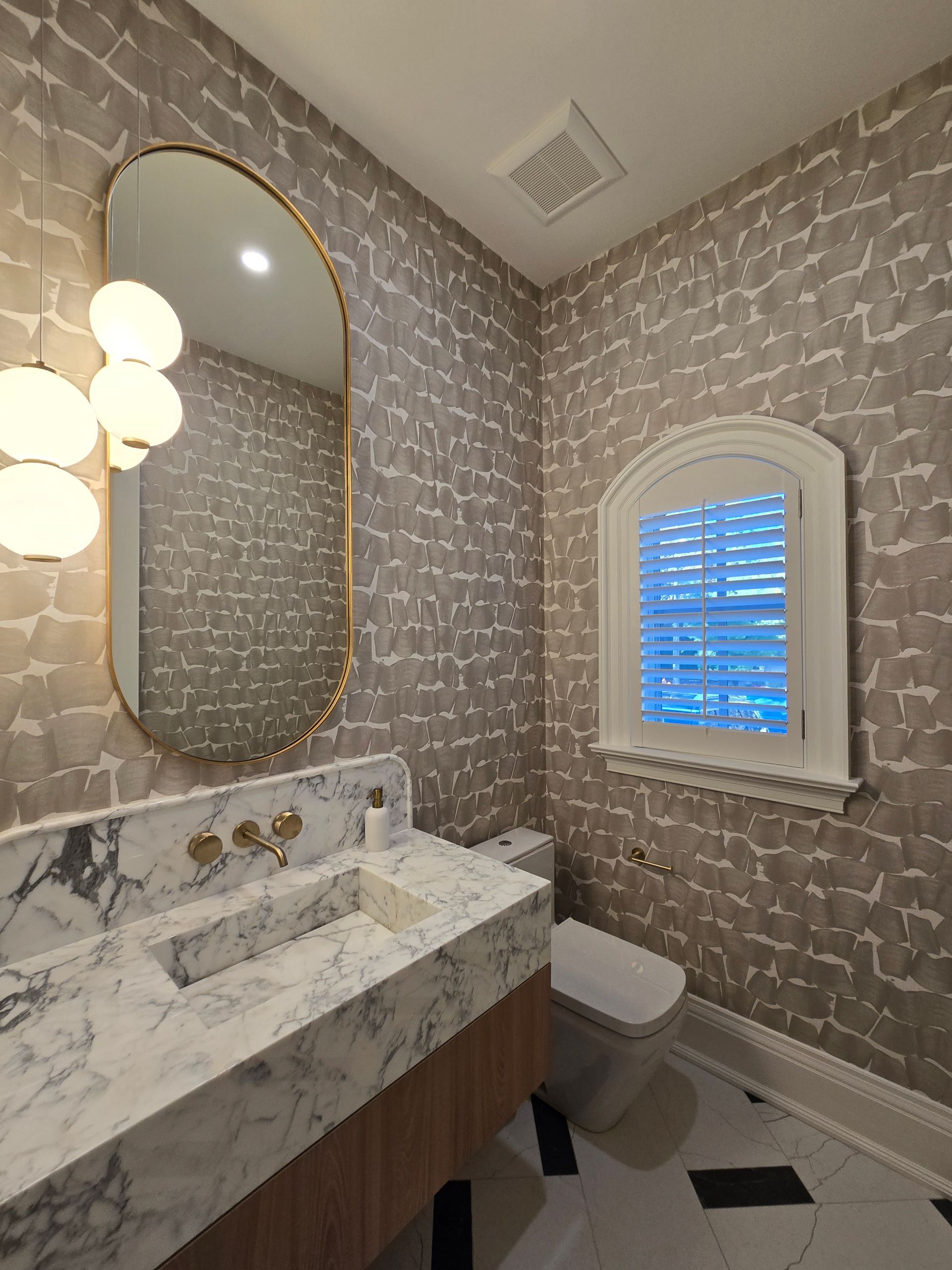 Powder room with marble vanity, gold mirror, patterned wallpaper, and globe pendant lights.