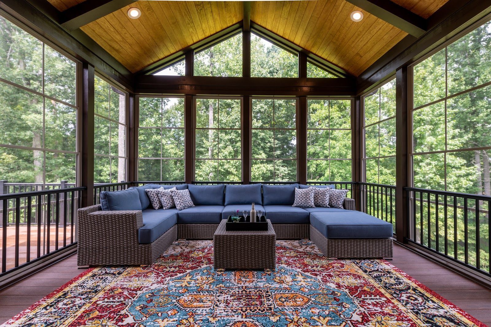 Screened-in porch with blue sectional, patterned rug, and brown wicker coffee table, overlooking trees.