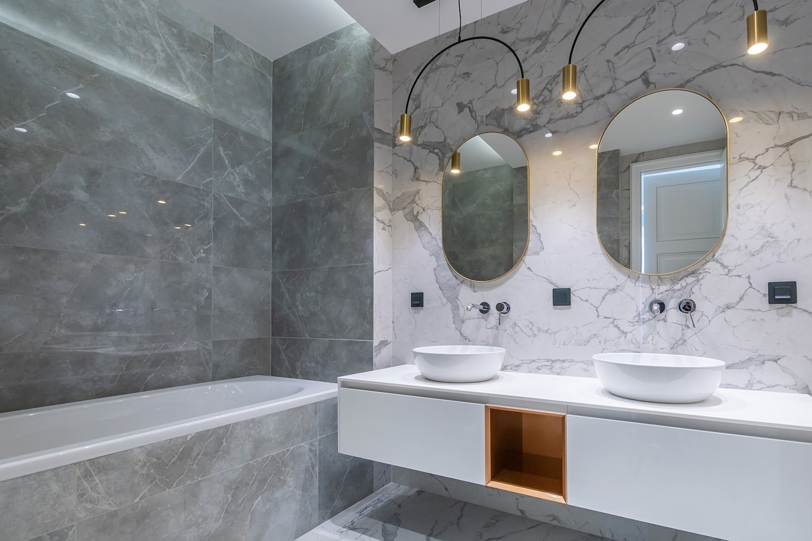 Modern bathroom with marble walls, two oval mirrors, two sinks, and a bathtub.