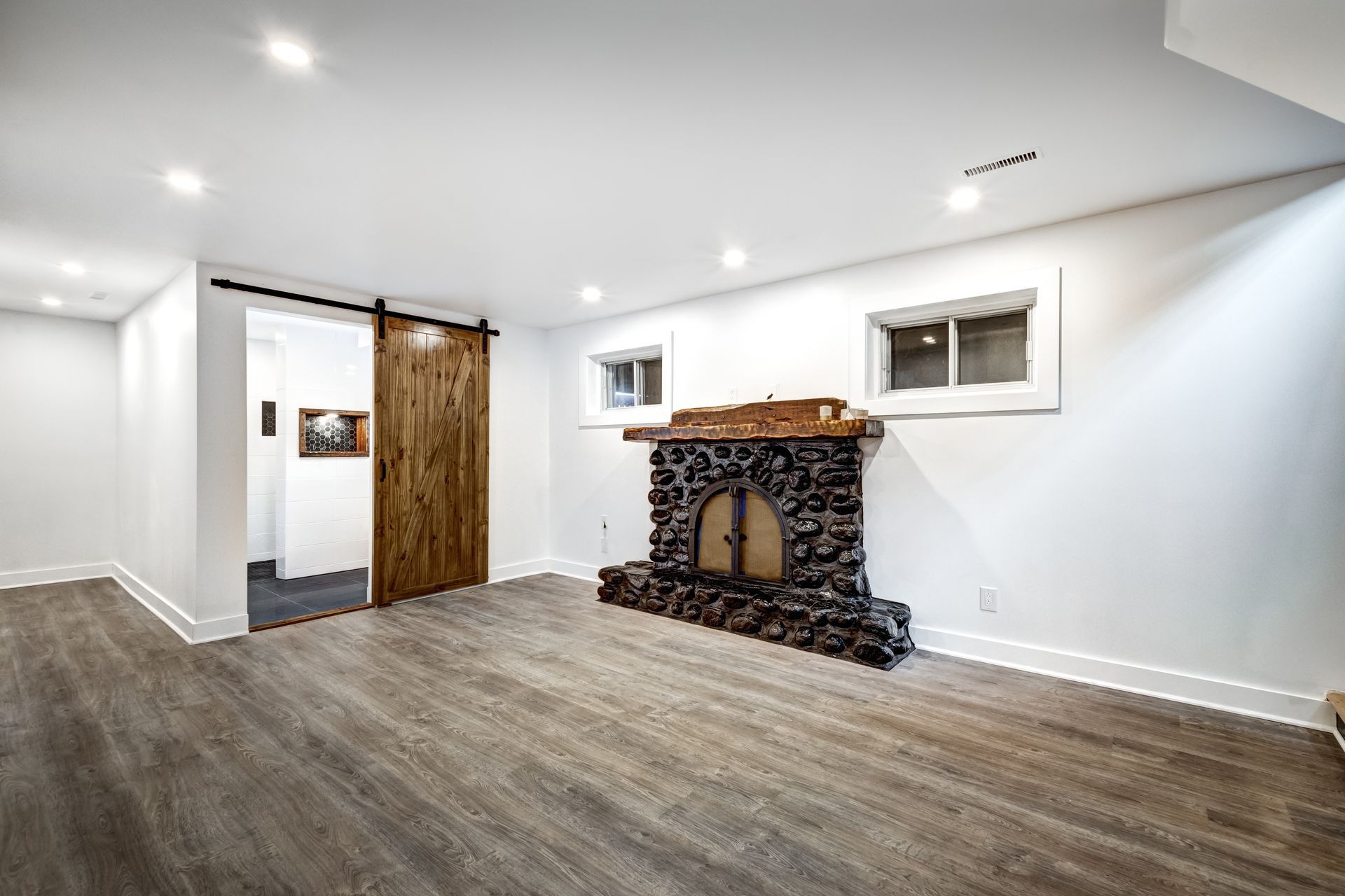 Basement room with a stone fireplace and wooden floor, a barn door, and small windows.