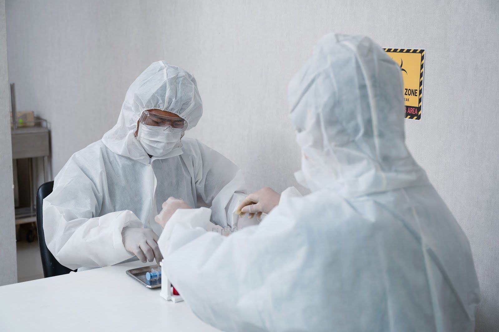 Two people in white protective suits inside a room, possibly a medical setting. One is working at a table.