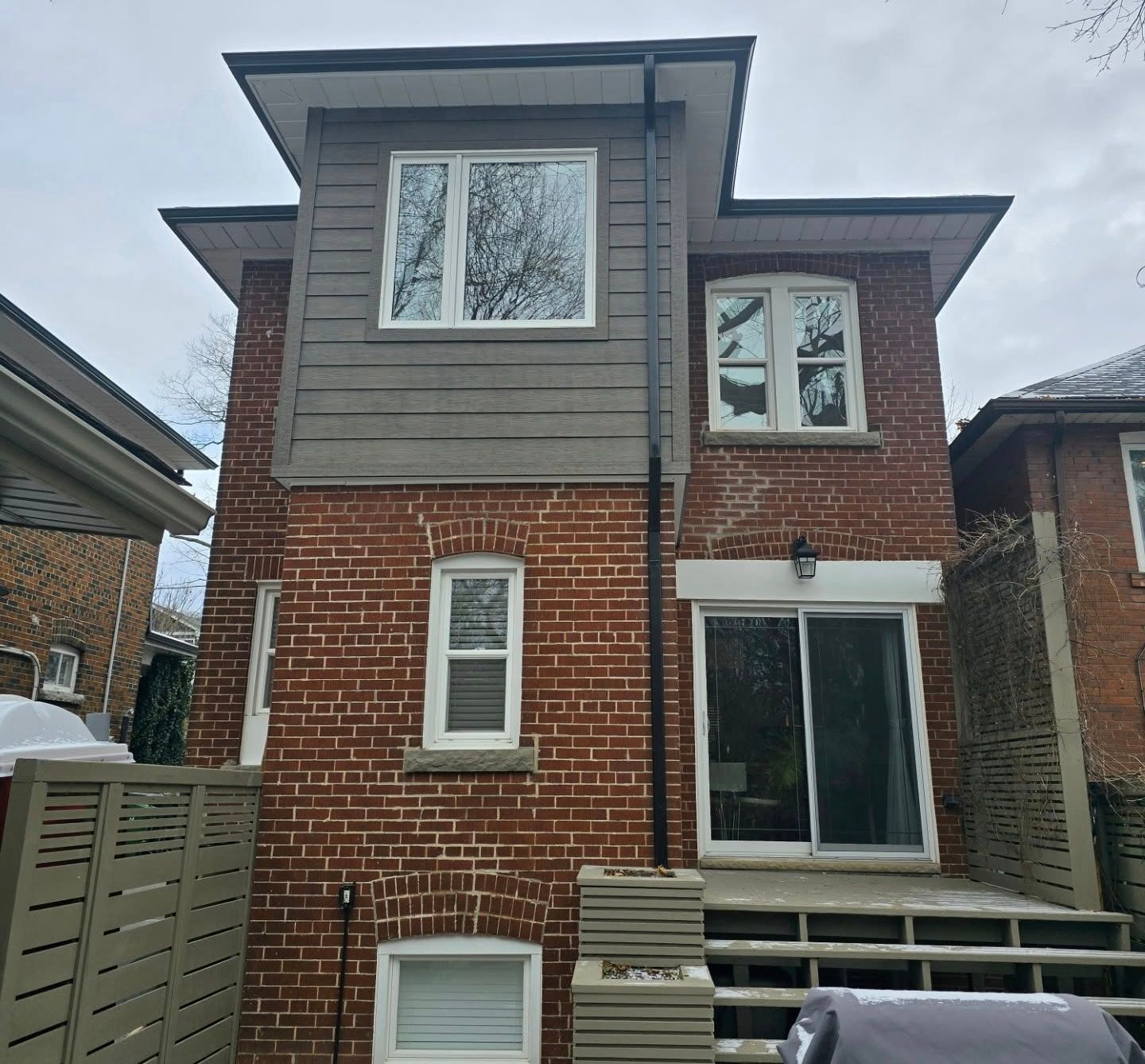 Red brick house with grey siding, white-framed windows, a black downspout, and a wooden deck.