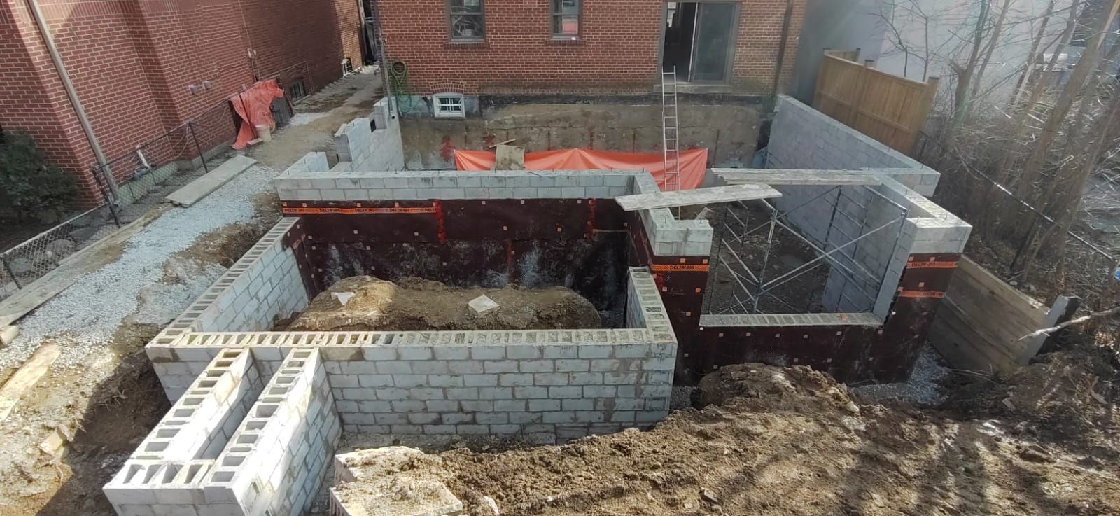 Construction site with concrete block foundation partially built, dirt, red brick building in background.