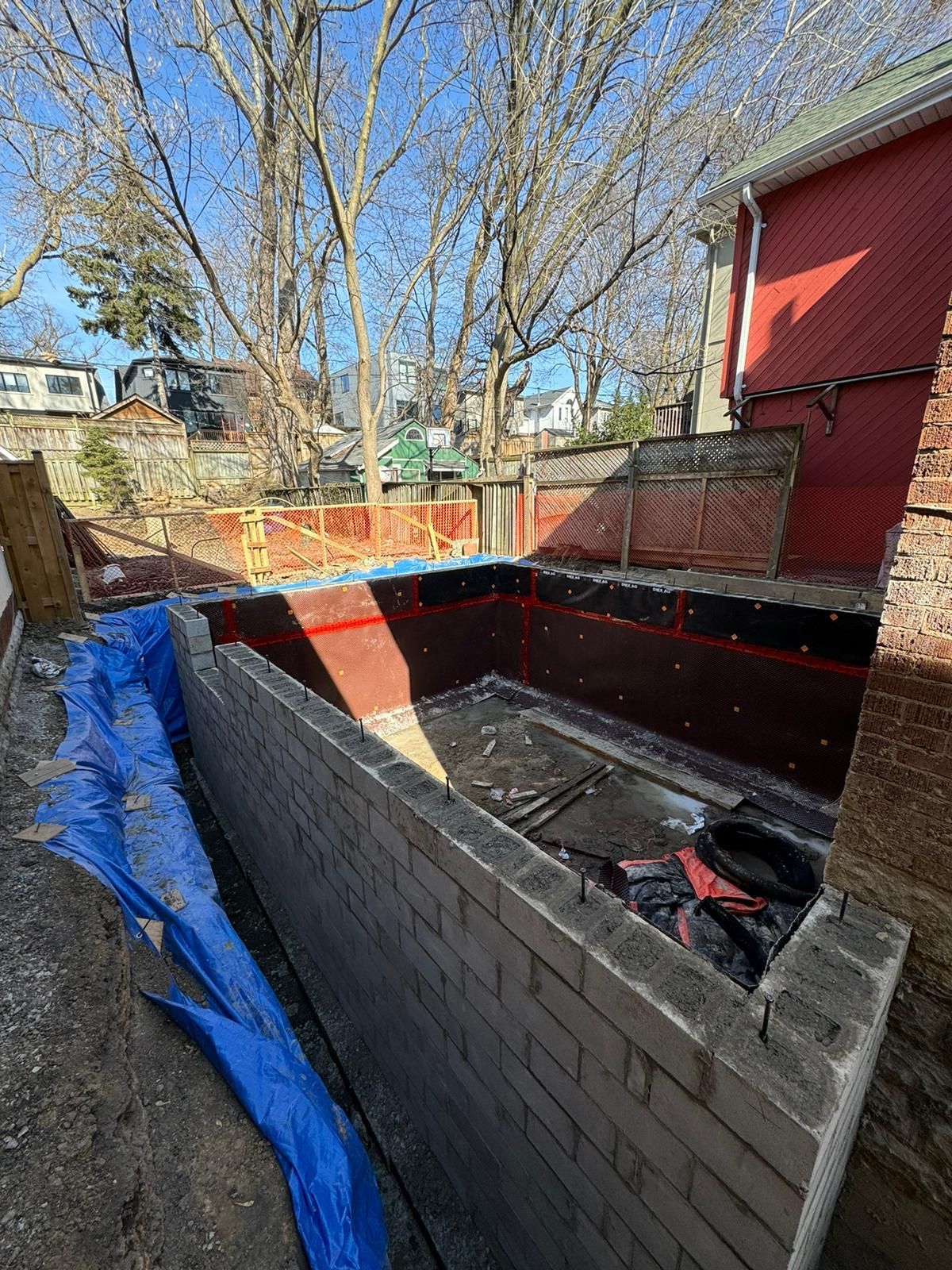 Construction site: brick foundation walls, lined with dark material, blue tarp, trees and red building in the background.
