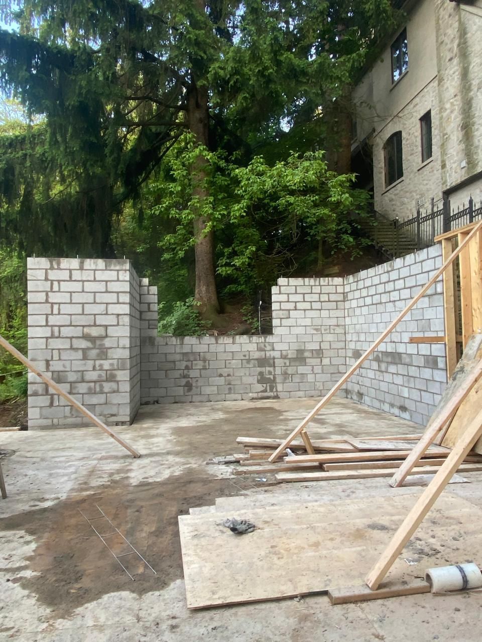 Construction site: cinder block walls, plywood flooring, wooden supports, trees, building in the background.