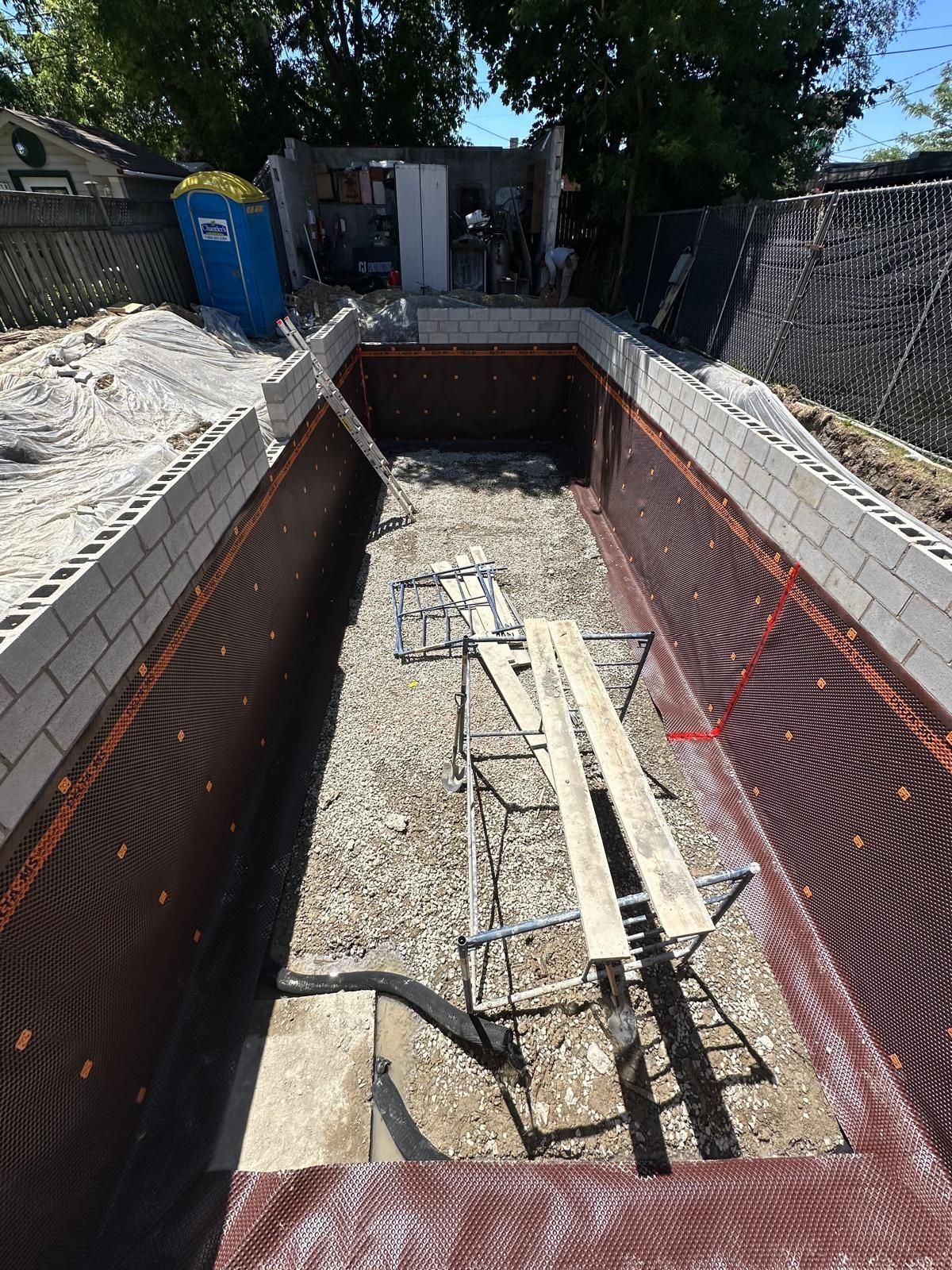 Pool under construction, rectangular shape. Dark brown liner, cinder block walls. Wooden planks on a frame.