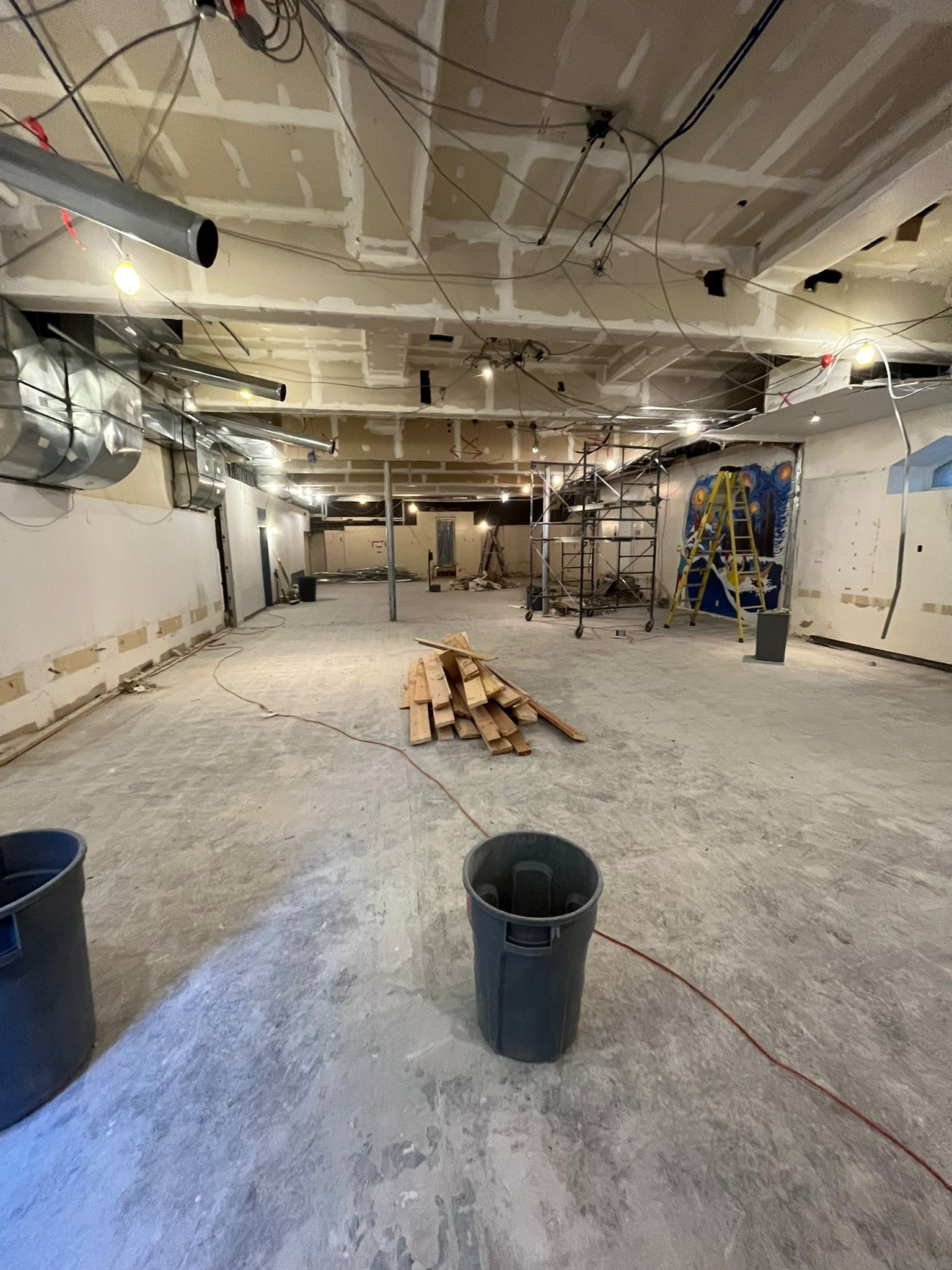 Interior construction site; exposed ceiling, concrete floor, drywall, debris, two trash cans.