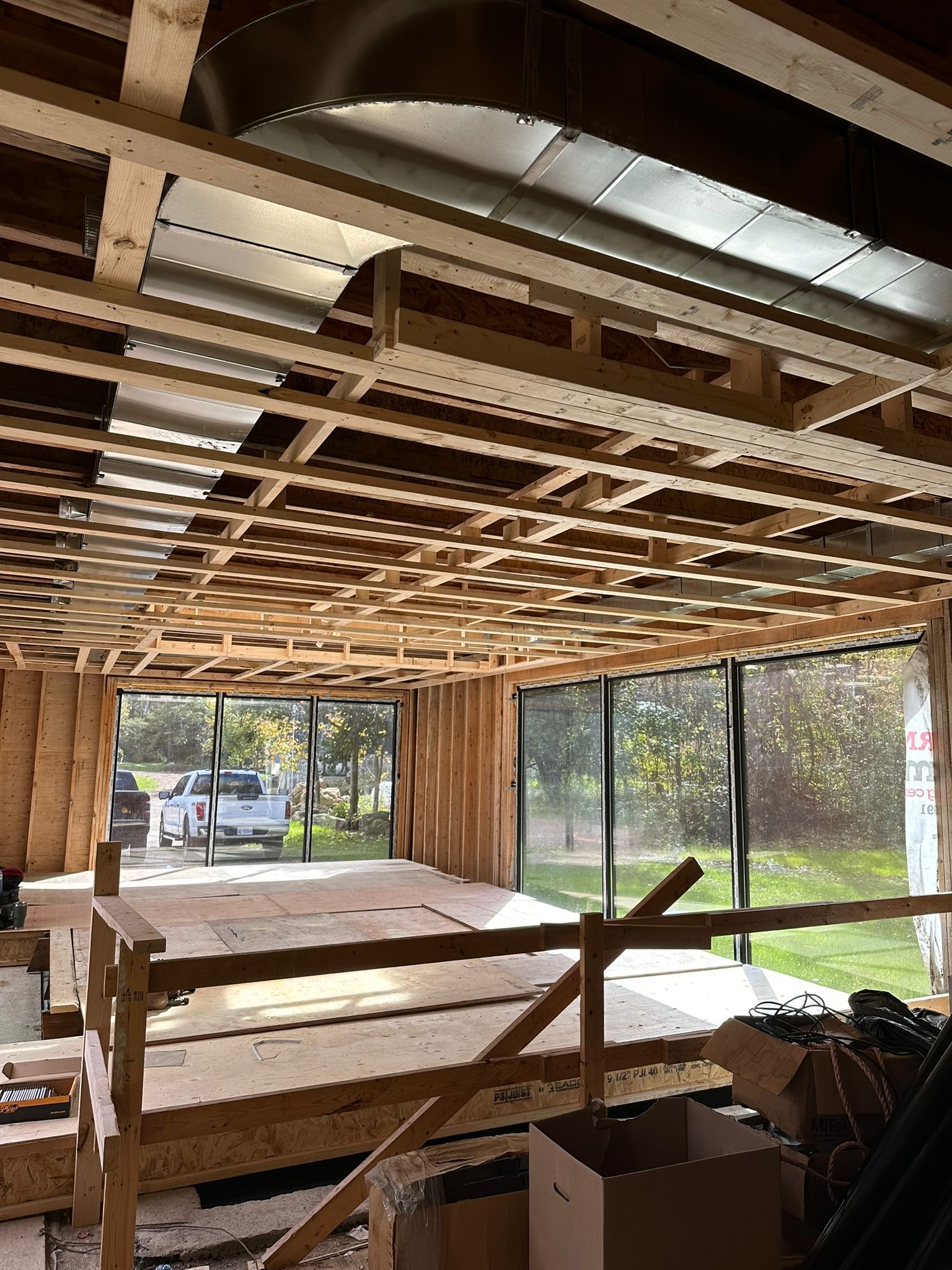 Interior view of a building under construction, showing wooden framing, ductwork, and large windows with outdoor view.
