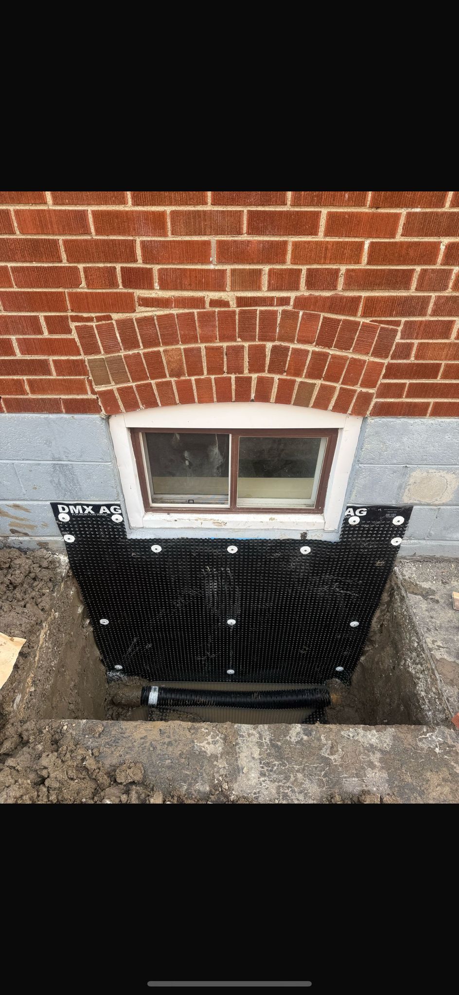 Exterior view of a brick house with a window well construction in progress, featuring black protective material.