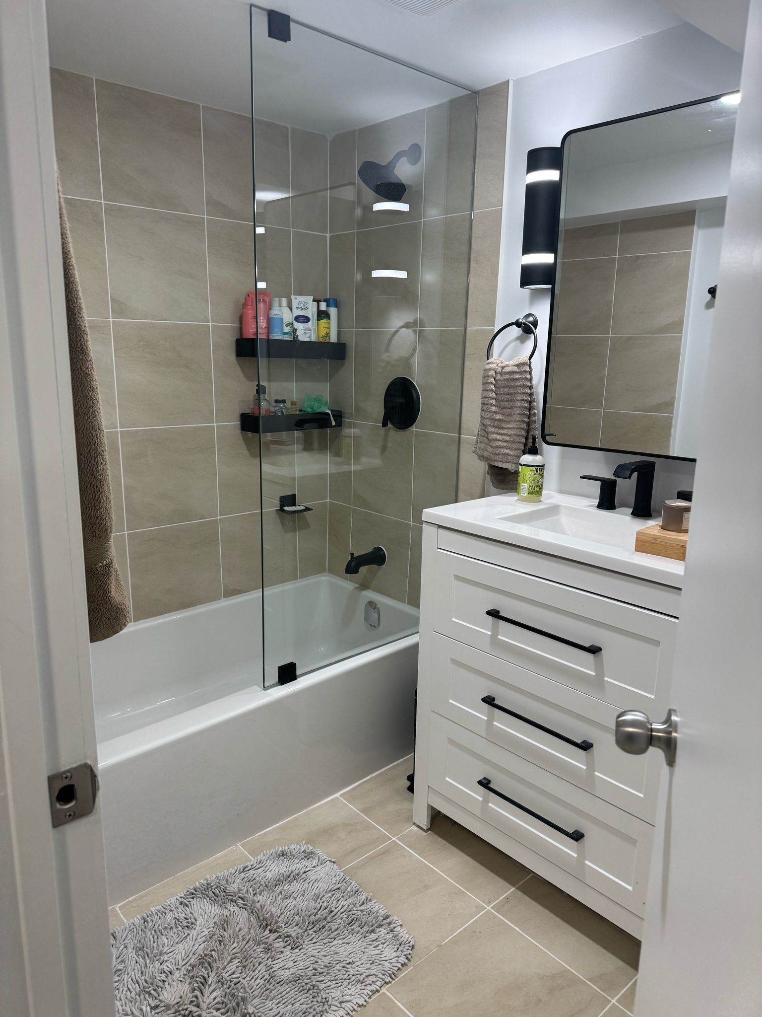 Modern bathroom with white vanity, bathtub, glass shower screen, and tan tile walls.