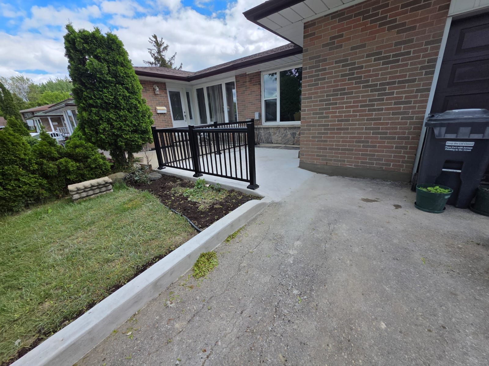 Black railing in front of a brick house with a concrete walkway and driveway. Green lawn in front.