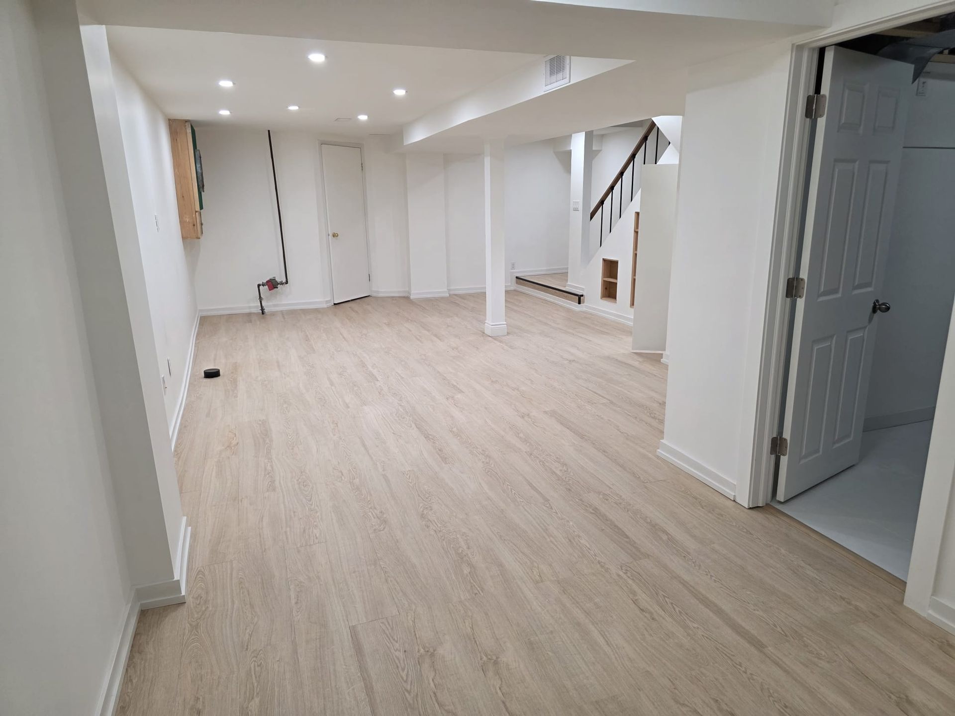 Empty, newly renovated basement with light wood-look flooring and white walls. Staircase, doors, and exposed support beams are visible.