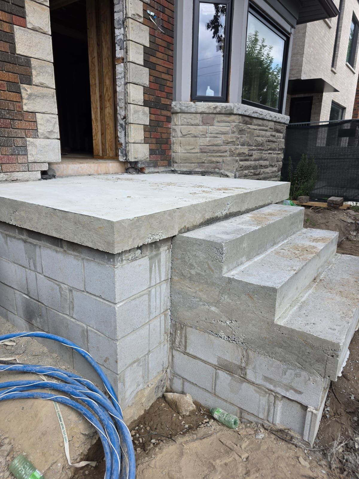 Concrete steps and porch under construction at a brick house. Blue hose on the ground.