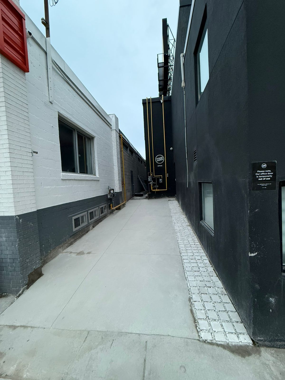 Alleyway between buildings, one white, one black. Concrete path slopes upward to a dark opening.