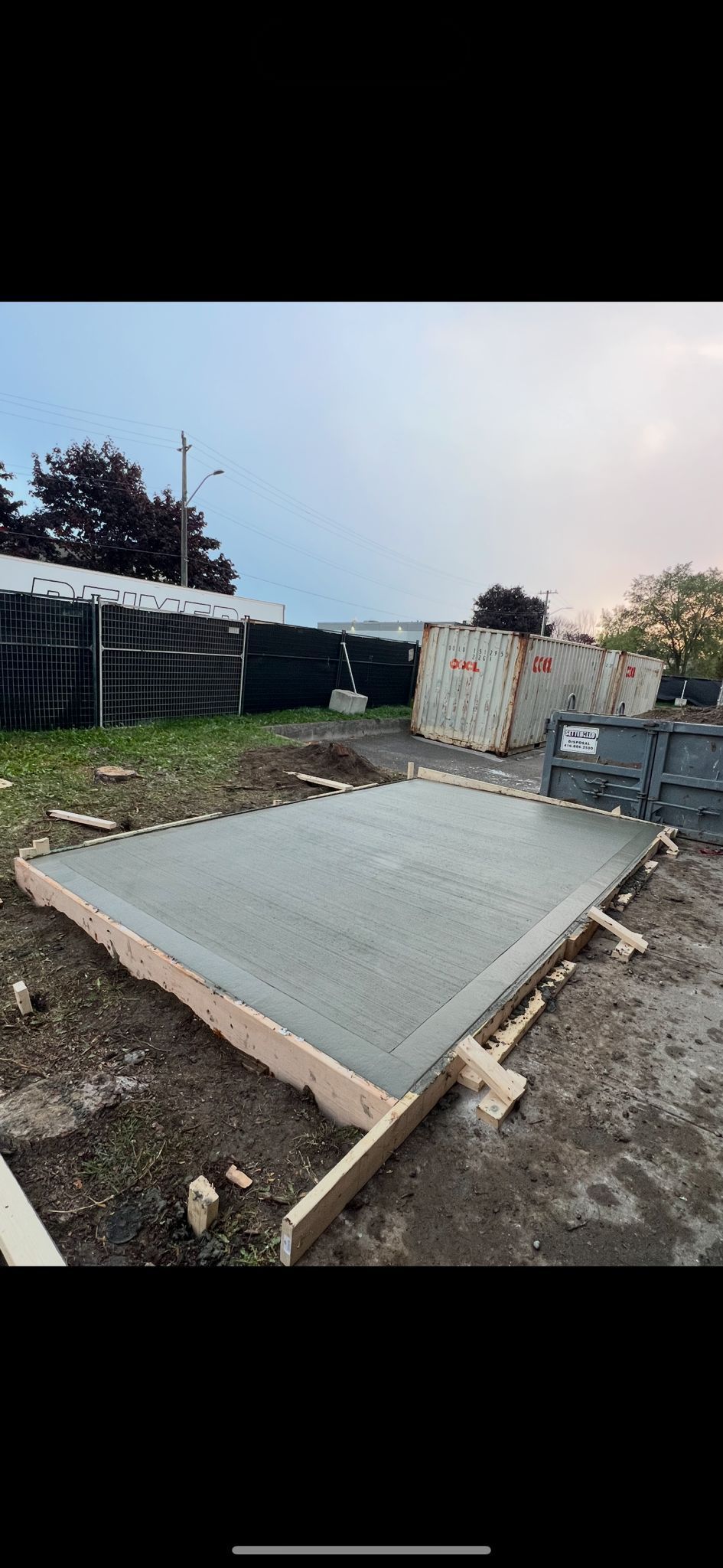 Freshly poured concrete slab with wooden frame, in an outdoor setting with green vegetation and overcast sky.