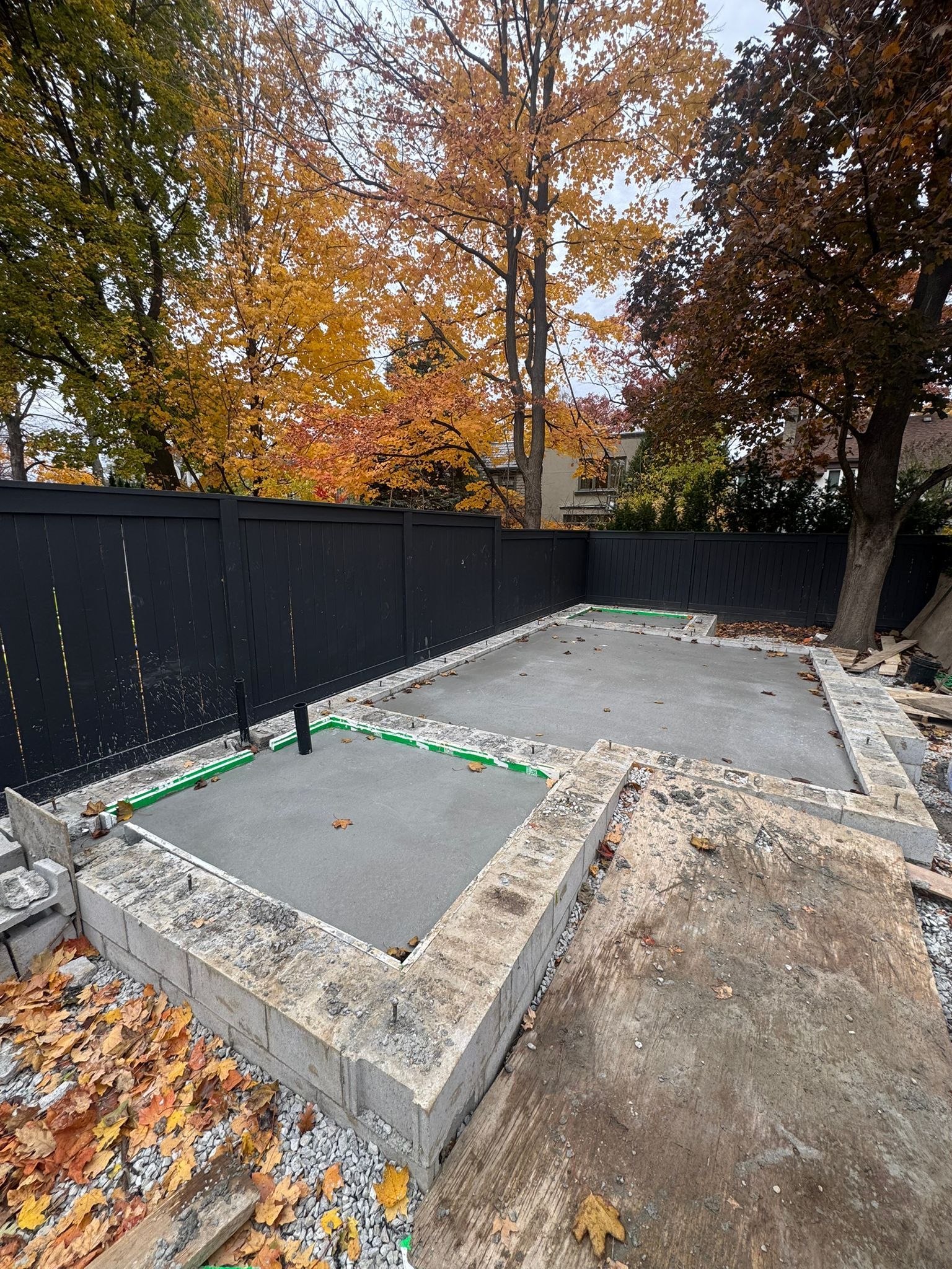 Concrete patio under construction, bordered by stone and black fence, with autumn foliage in background.