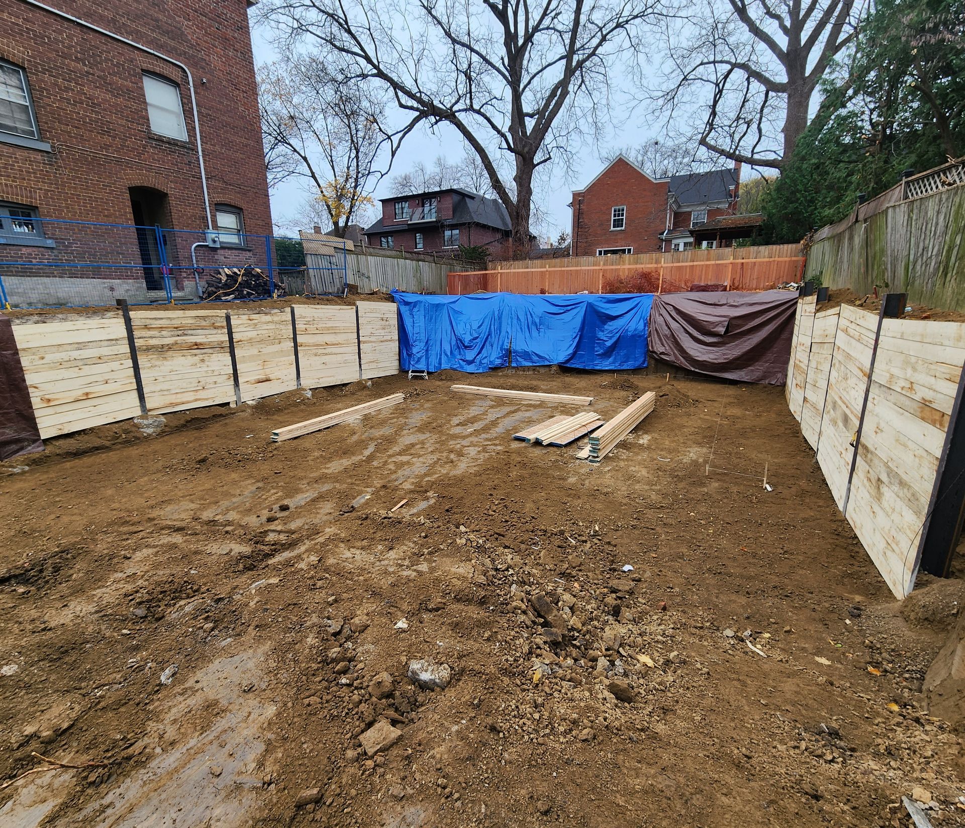 Construction site: wooden retaining walls, dirt ground, blue and brown tarps, brick building in the background.