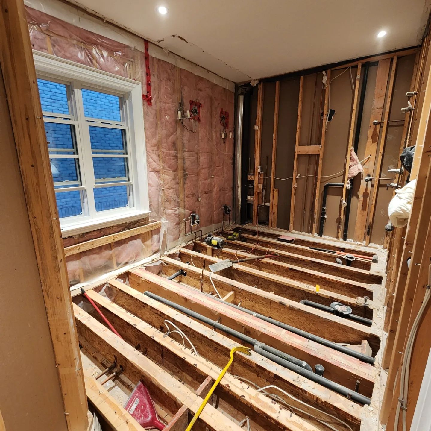 Bathroom under renovation, with exposed wooden framing, insulation, plumbing, and a window.