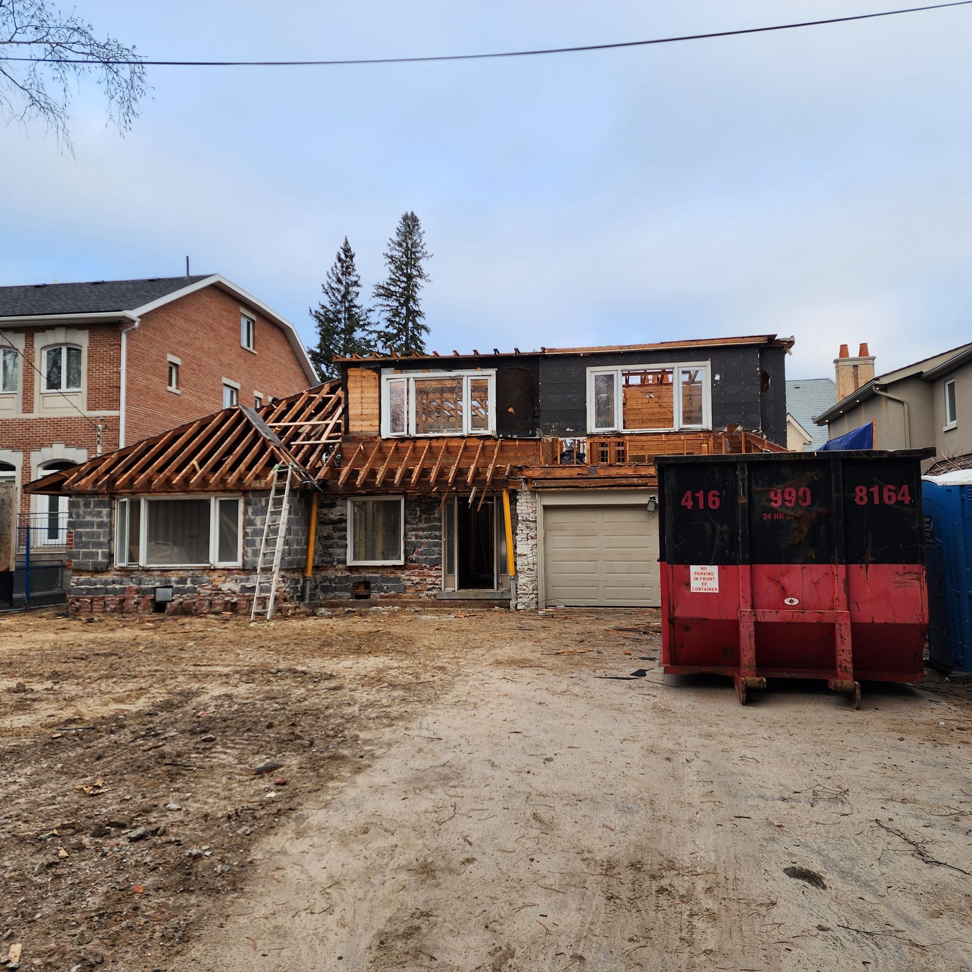 House under construction; roof partially removed, debris on ground, red dumpster, overcast sky.