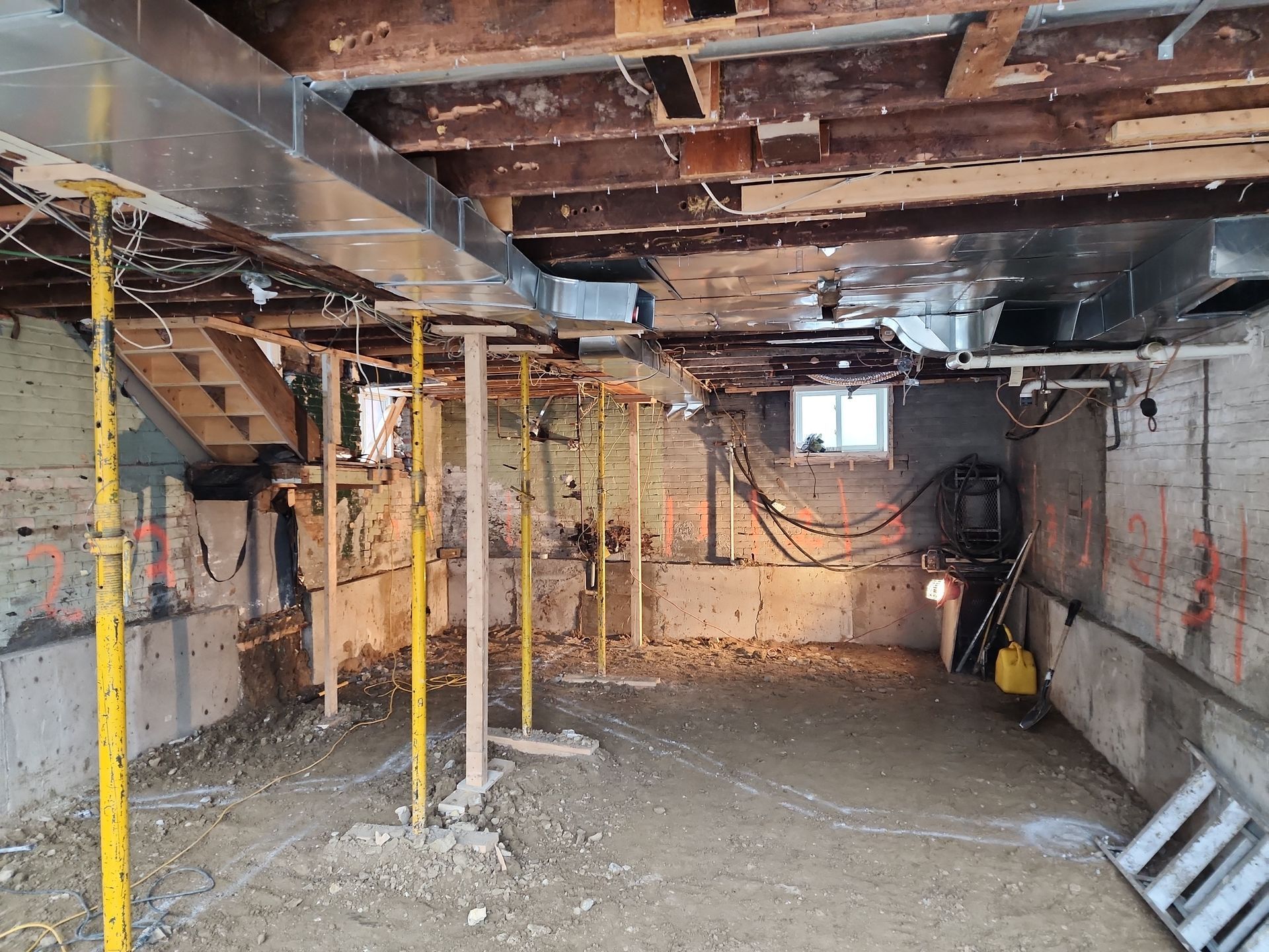 Basement under construction, exposed beams, ductwork, supporting jacks, dirt floor, and concrete walls.