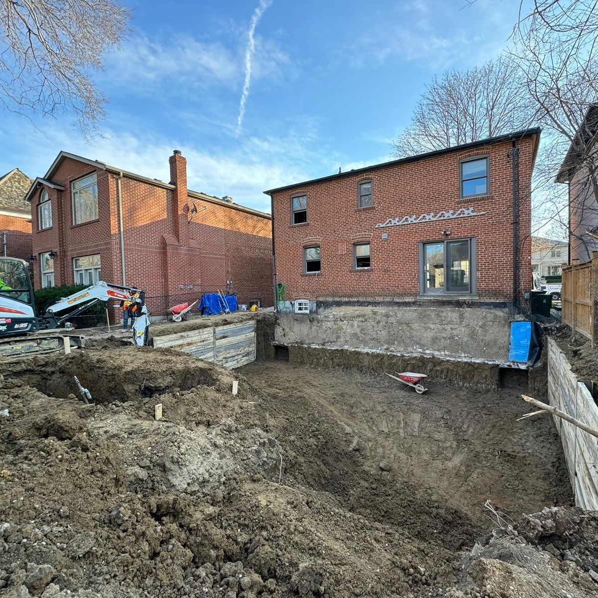 Construction site with two brick houses; excavation in progress with retaining wall and backhoe.