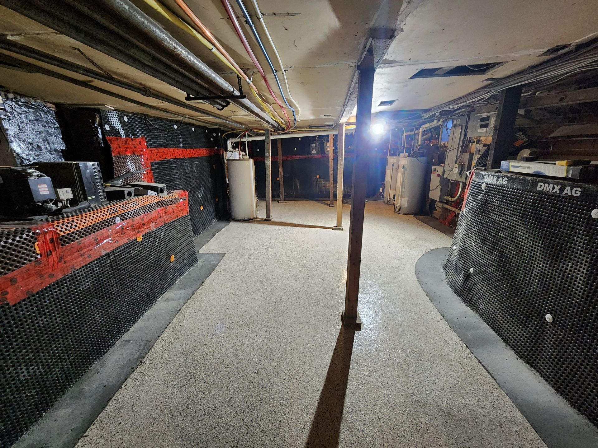 Basement interior with exposed pipes and black patterned walls, concrete floor, central support beams.