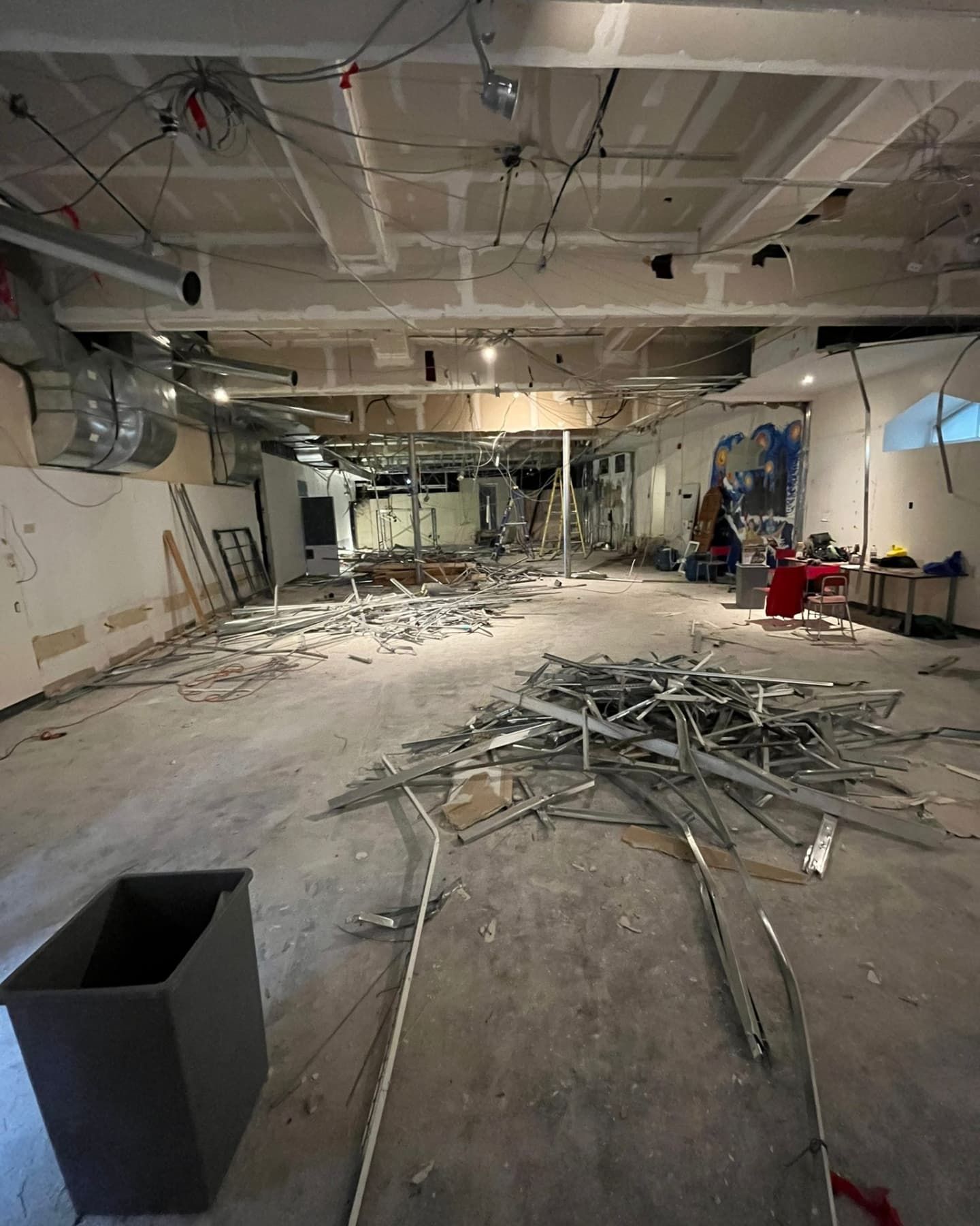 Interior demolition scene; debris scattered on concrete floor. Exposed ceiling beams, wiring, and ductwork visible.