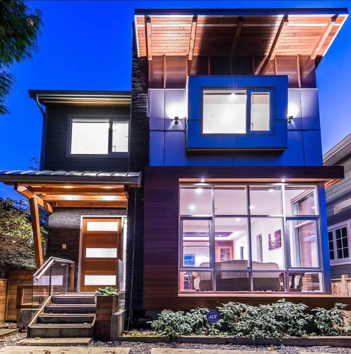 Modern two-story house with wood and blue paneling, large windows, and an illuminated entryway.