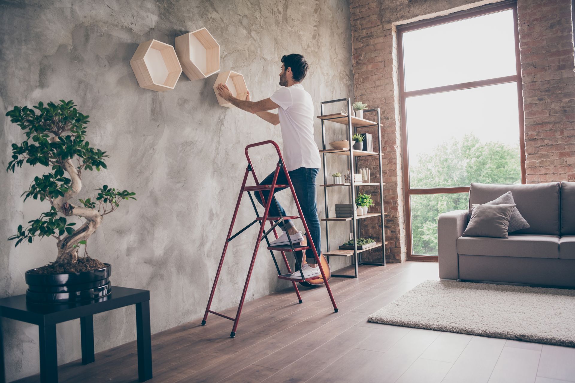 A man is standing on a ladder in a living room hanging shelves on a wall.