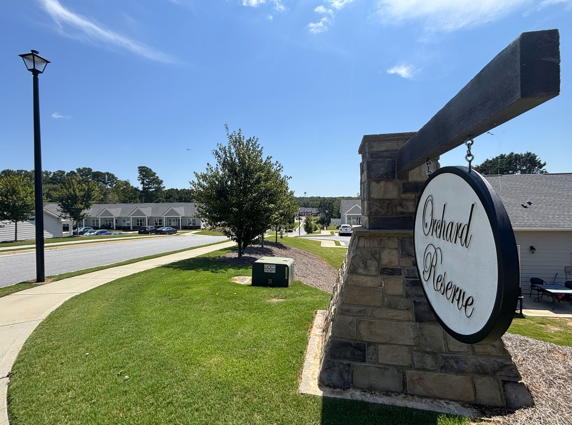 Stone entrance sign at a suburban complex under a bright blue sky, with lawns, trees, and parked cars nearby