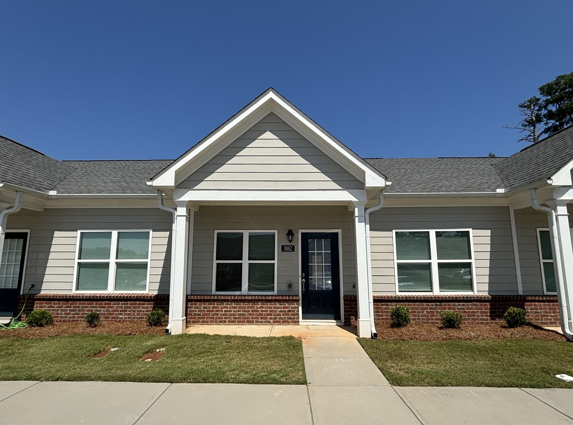 Single-story beige house with white trim, brick accents, and a front walkway under a clear blue sky