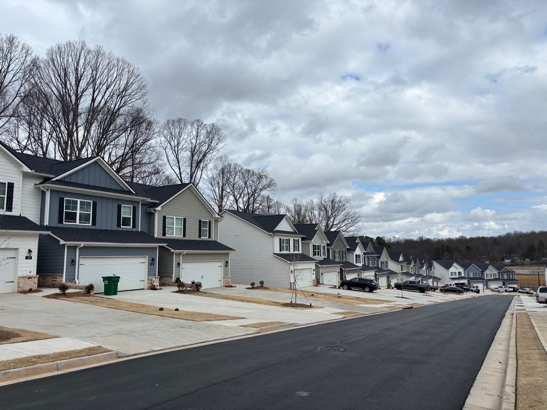 Suburban street with new houses, driveways, and leafless trees under a cloudy sky
