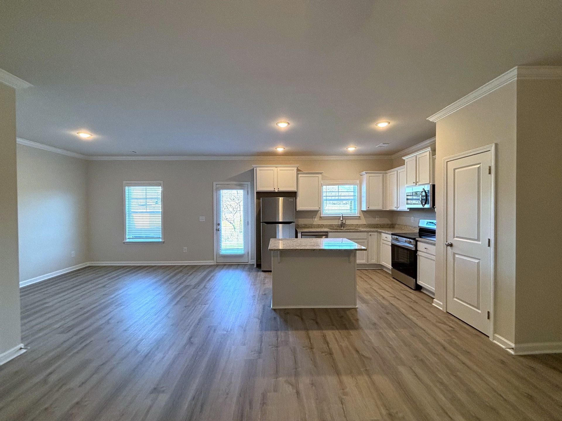 Empty open-plan kitchen and living area with light wood floors, white cabinets, and recessed ceiling lights.