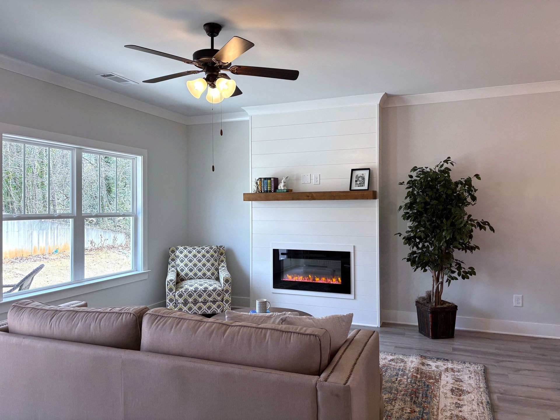 Bright living room with beige sofa, fireplace, large window, ceiling fan, and potted plant.