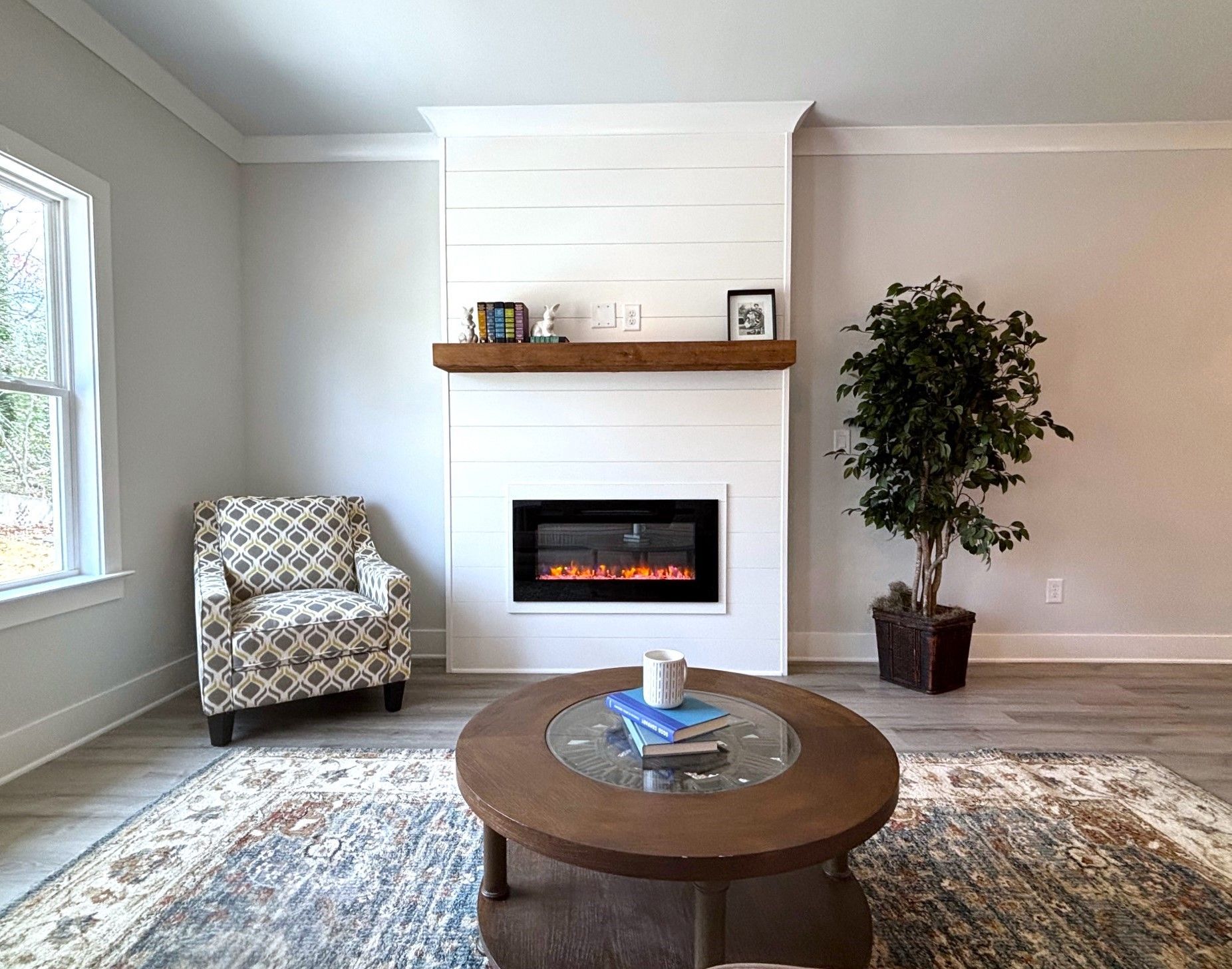 Bright living room with fireplace, armchair, coffee table, rug, and potted tree beside a window